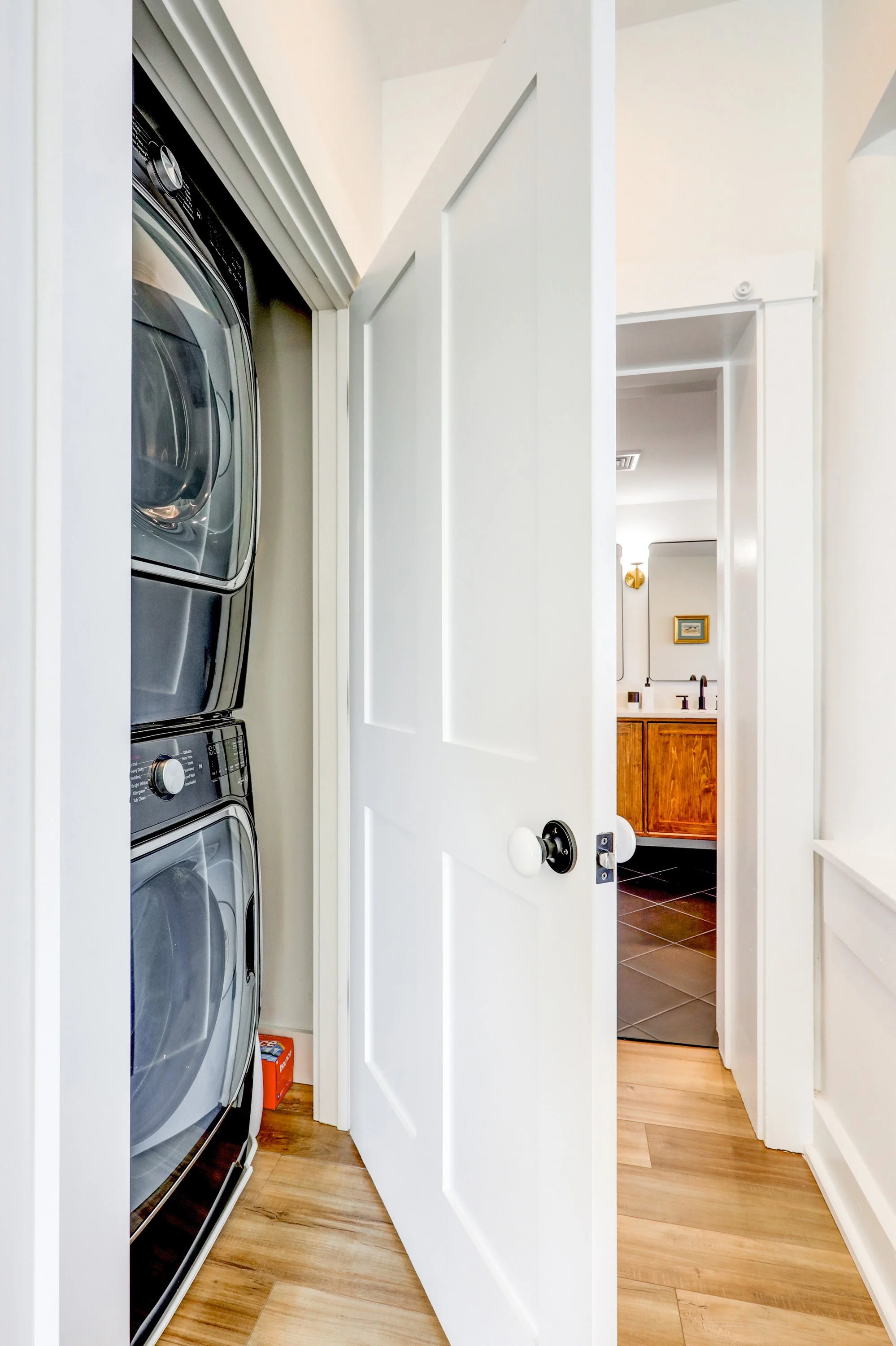 Small laundry area with stacked silver washing machine and dryer, white closet door partially open, and a glimpse into a bathroom with wooden vanity and black countertop.