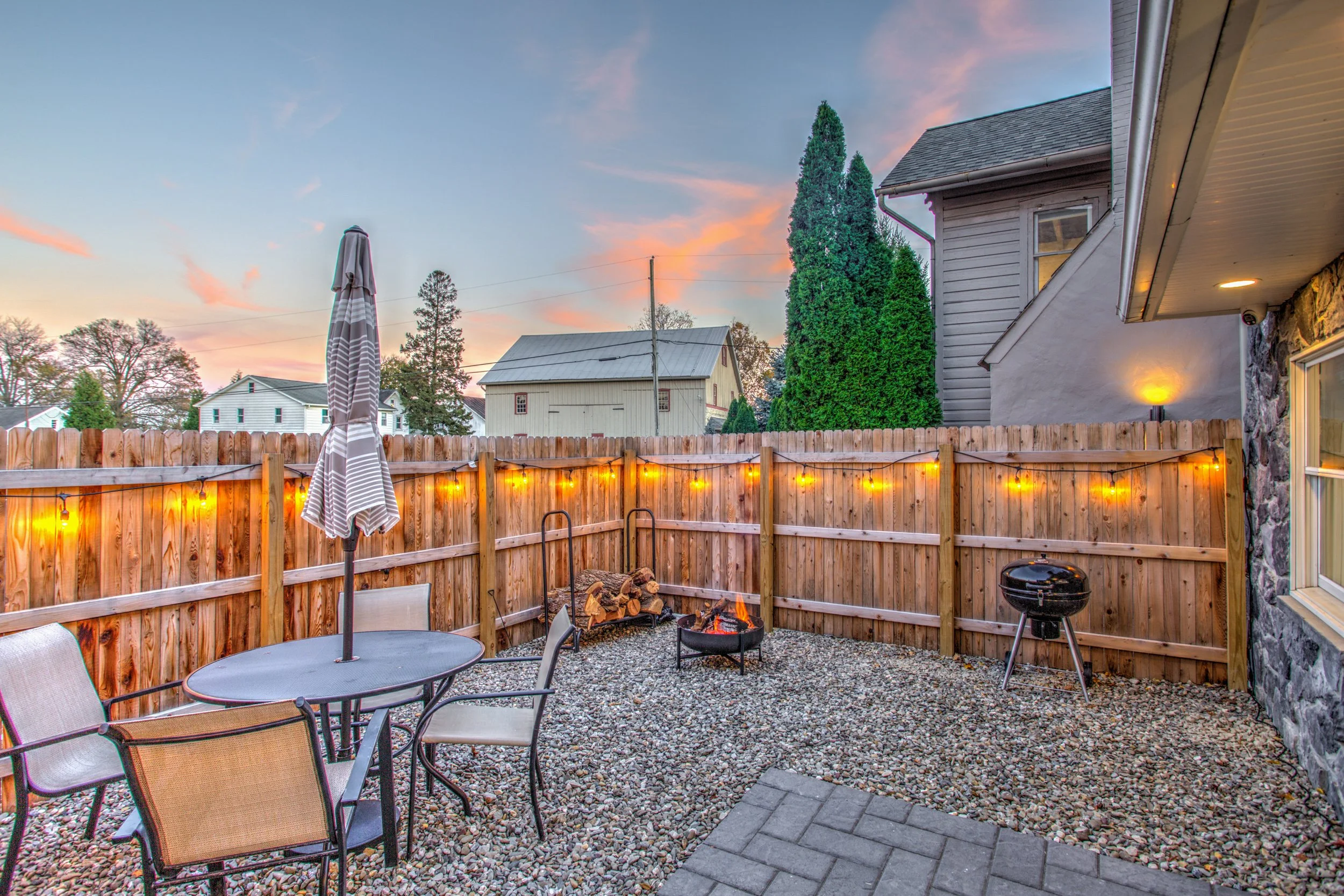 Backyard patio with a gravel surface, outdoor dining table and chairs, a closed umbrella, a small fire pit with logs, string lights on a wooden fence, and a view of neighboring houses and tall trees during sunset.