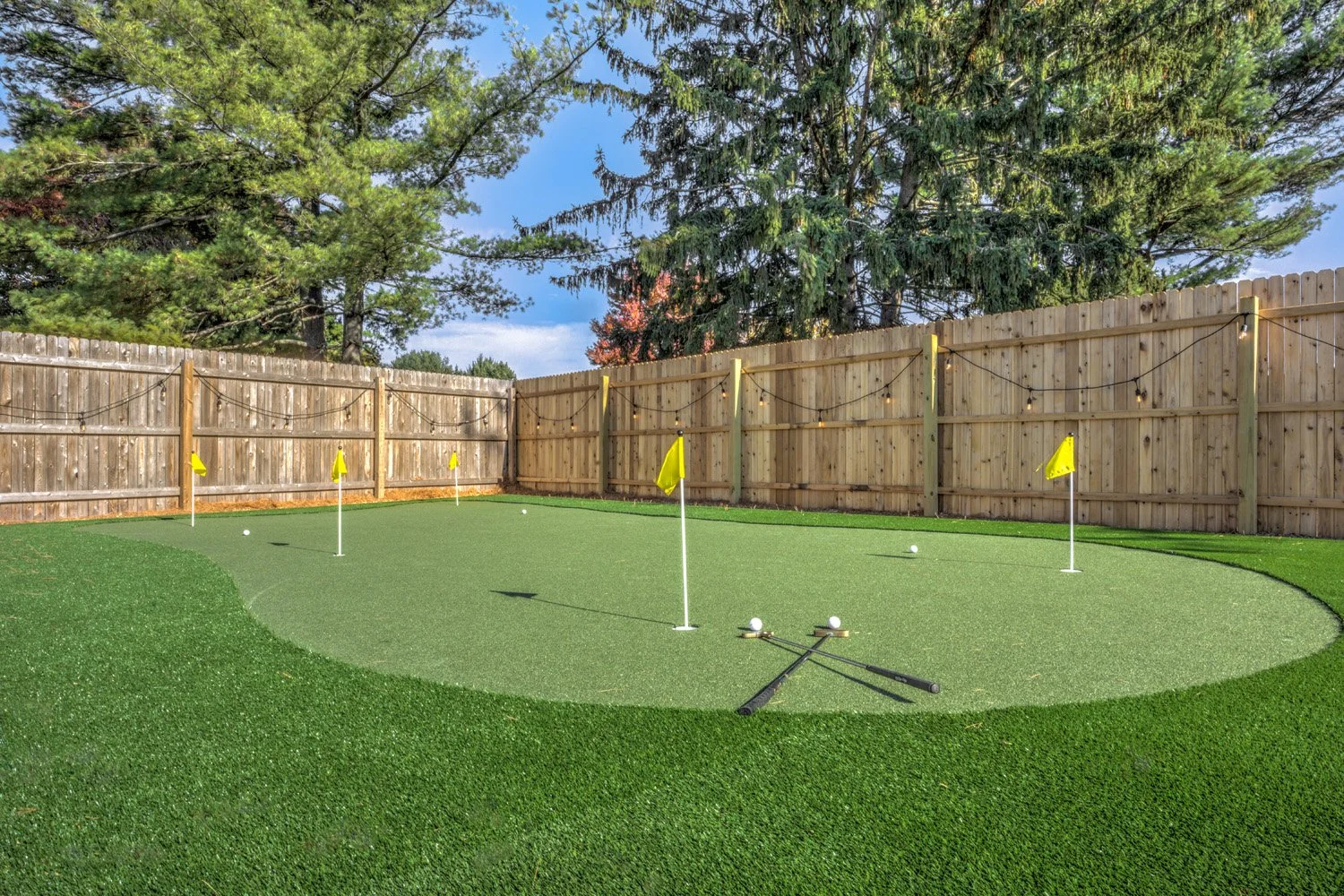 A backyard putting green with four golf holes marked by yellow flags, surrounded by a wooden fence and trees, with two golf practice putters lying on the green.