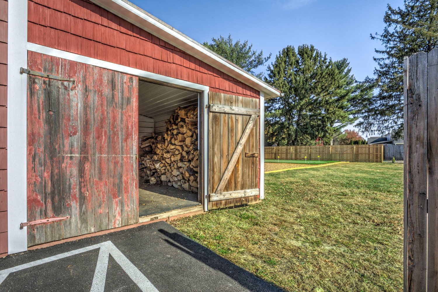 Wooden shed with open door revealing stored firewood, located on a paved area with parking lines, surrounded by a grassy yard with trees and a wooden fence in the background.