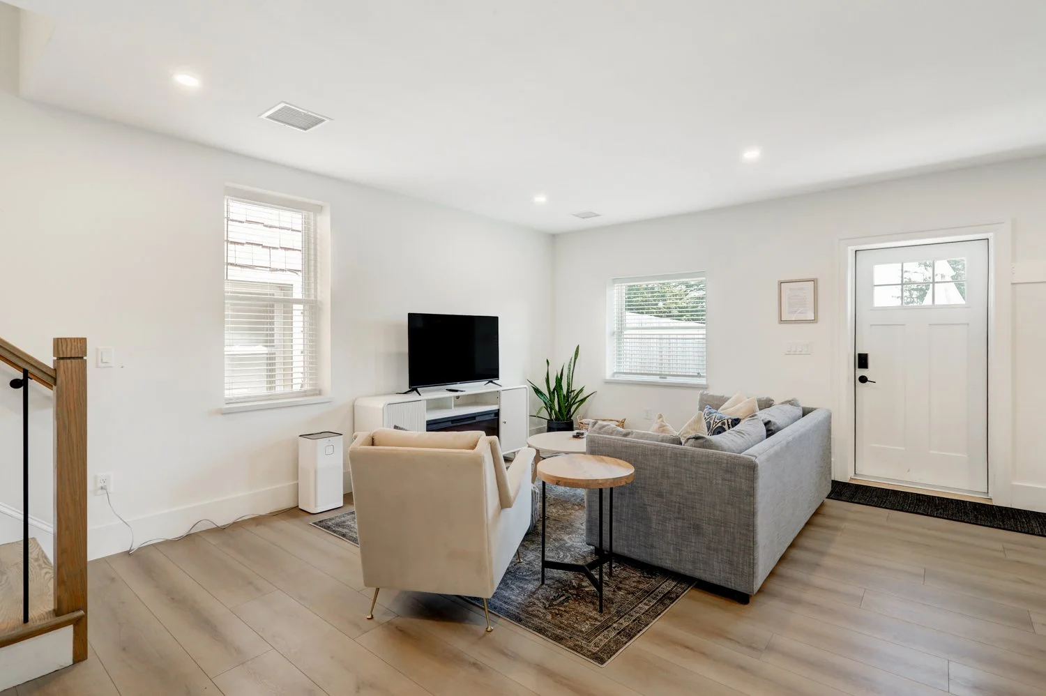 Modern living room with gray and beige furniture, a flat-screen TV, wooden floors, and large windows with white blinds.