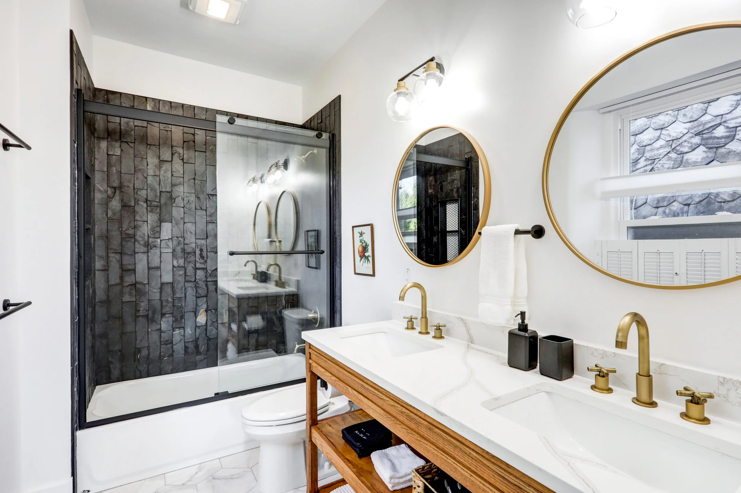 Modern bathroom with white walls, marble countertops, gold fixtures, a wooden vanity with open shelving, and a large round mirror. There is a black soap dispenser and black toothbrush holder on the counter, a towel hanging on a black towel bar, and a