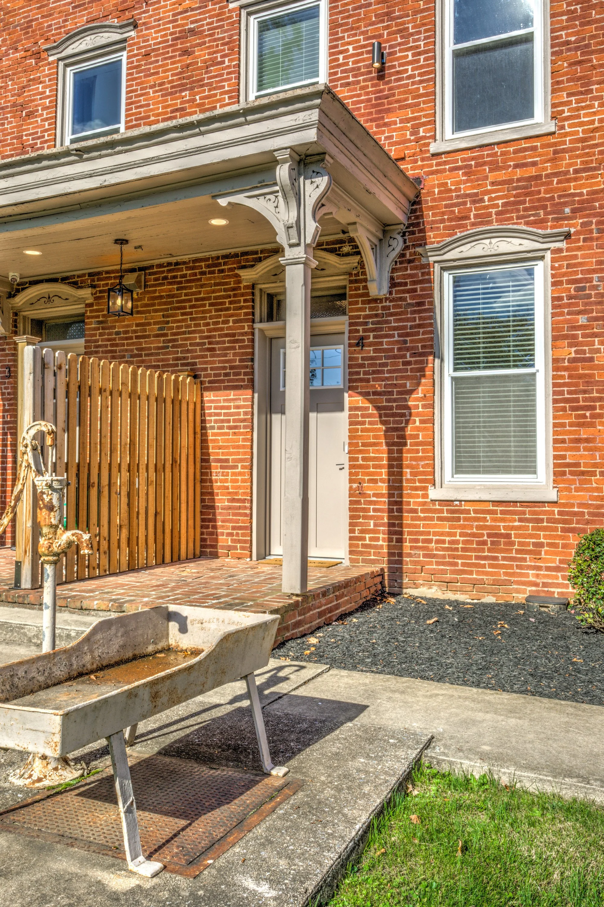 A brick house with a small porch area and large windows. There is an old, rusty wheelbarrow in the foreground, and a wooden fence near the entrance.