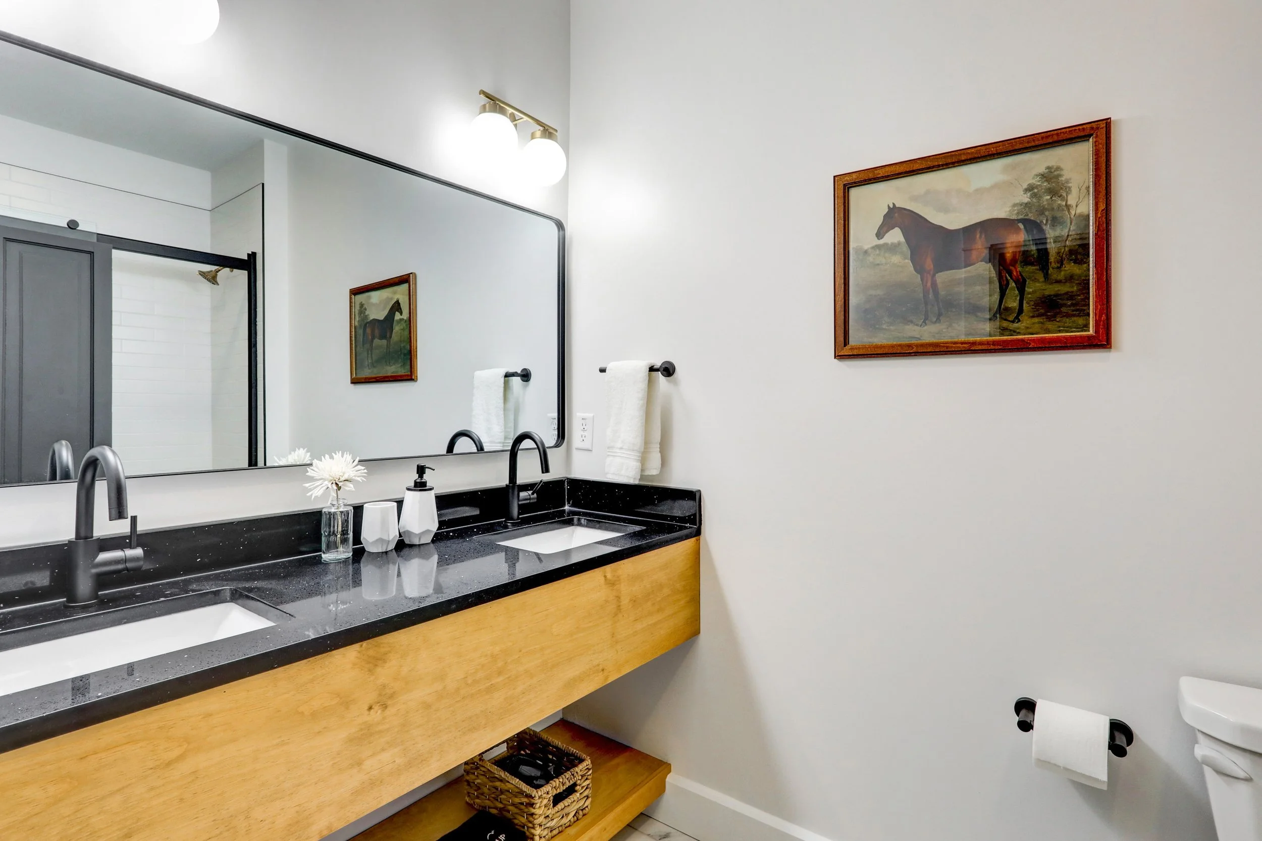 Modern bathroom with white walls, a large mirror, black countertop with a sink, and framed horse paintings on the wall.
