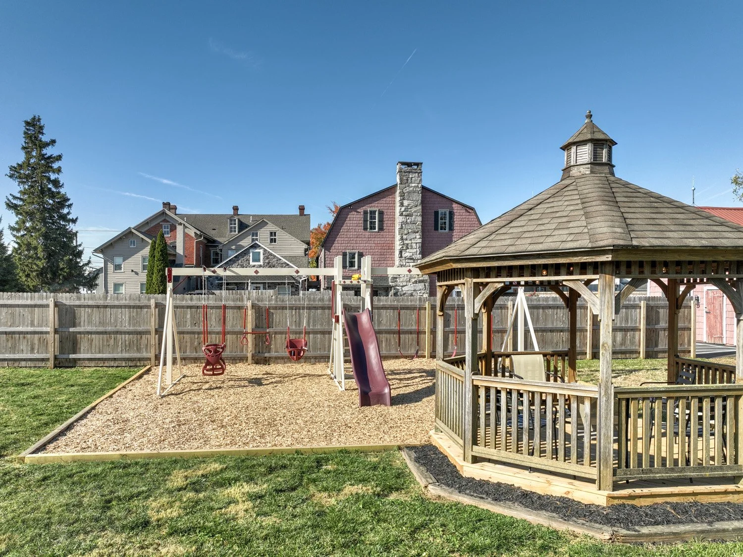 Child's backyard playground with swings, slide, sandbox, wooden gazebo, green grass, and houses in the background under a blue sky.