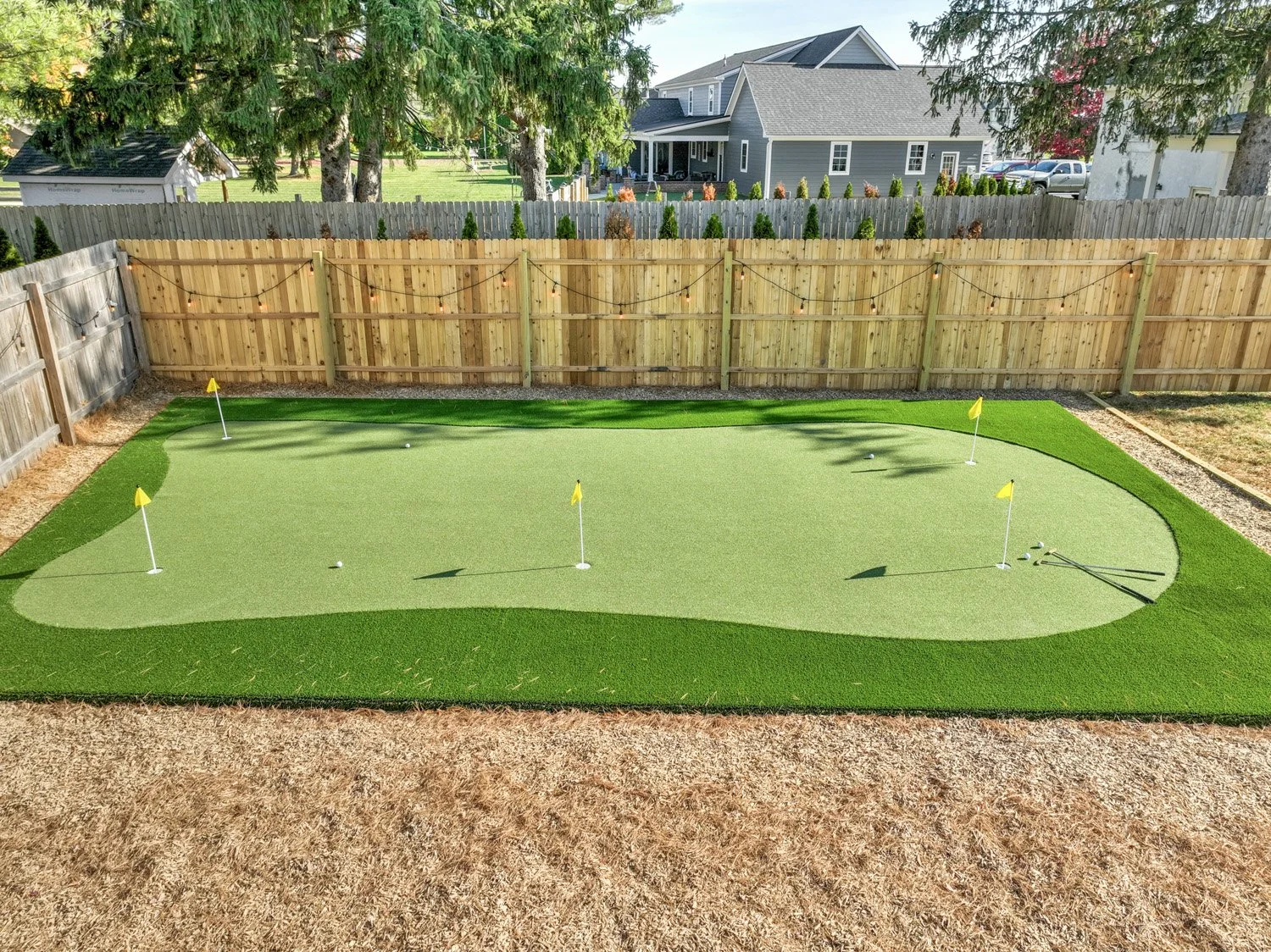 A backyard putting green with flags and golf balls, enclosed by a wooden fence, with trees and houses in the background.