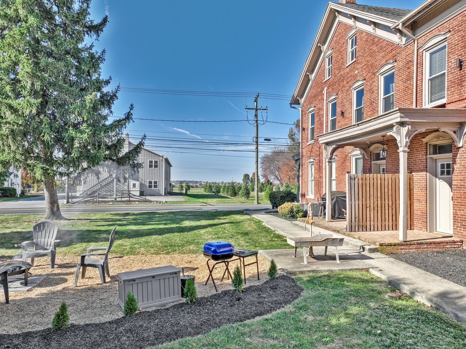 View of a backyard with chairs, a small grill, a lawn, a sidewalk, and a red brick building in the background on a clear, sunny day.