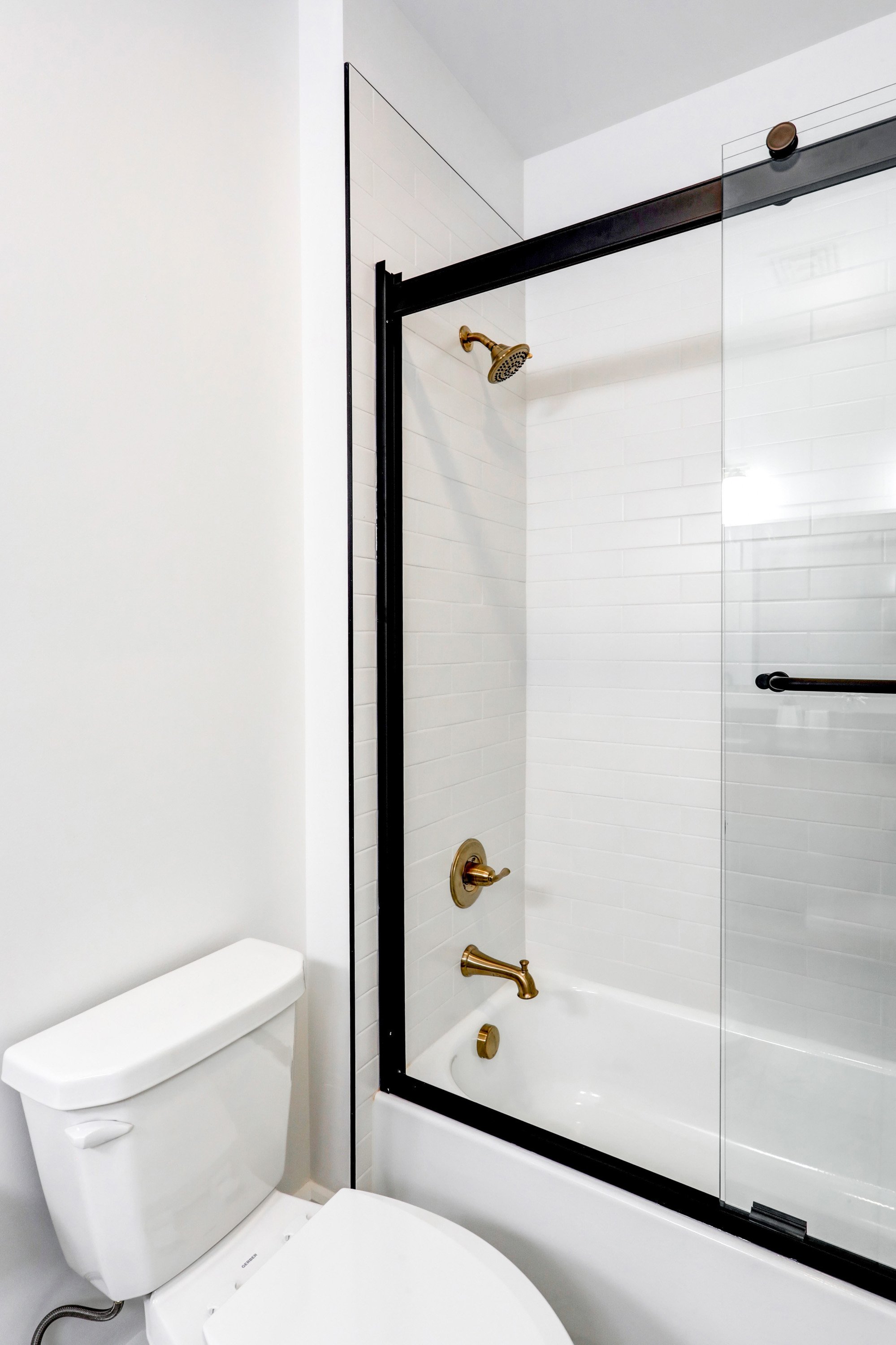 Bathroom with bathtub, glass shower door, white tiled wall, gold fixtures, and toilet.