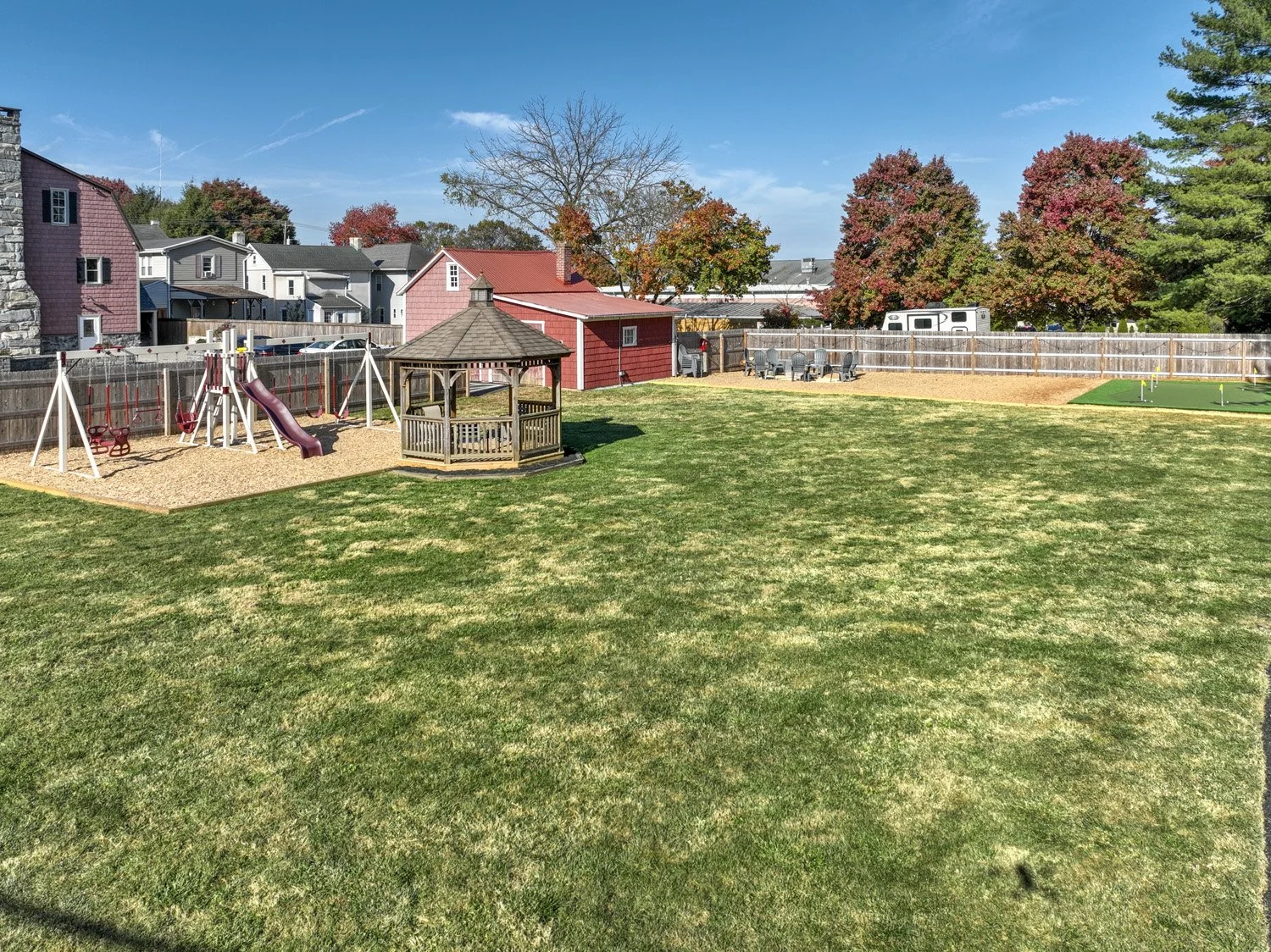 A backyard with a grassy lawn, a wooden gazebo, a sandbox with swings and a slide, a table with chairs, and a small putting green area enclosed by wooden fencing. Trees with autumn foliage are visible in the background under a clear blue sky.