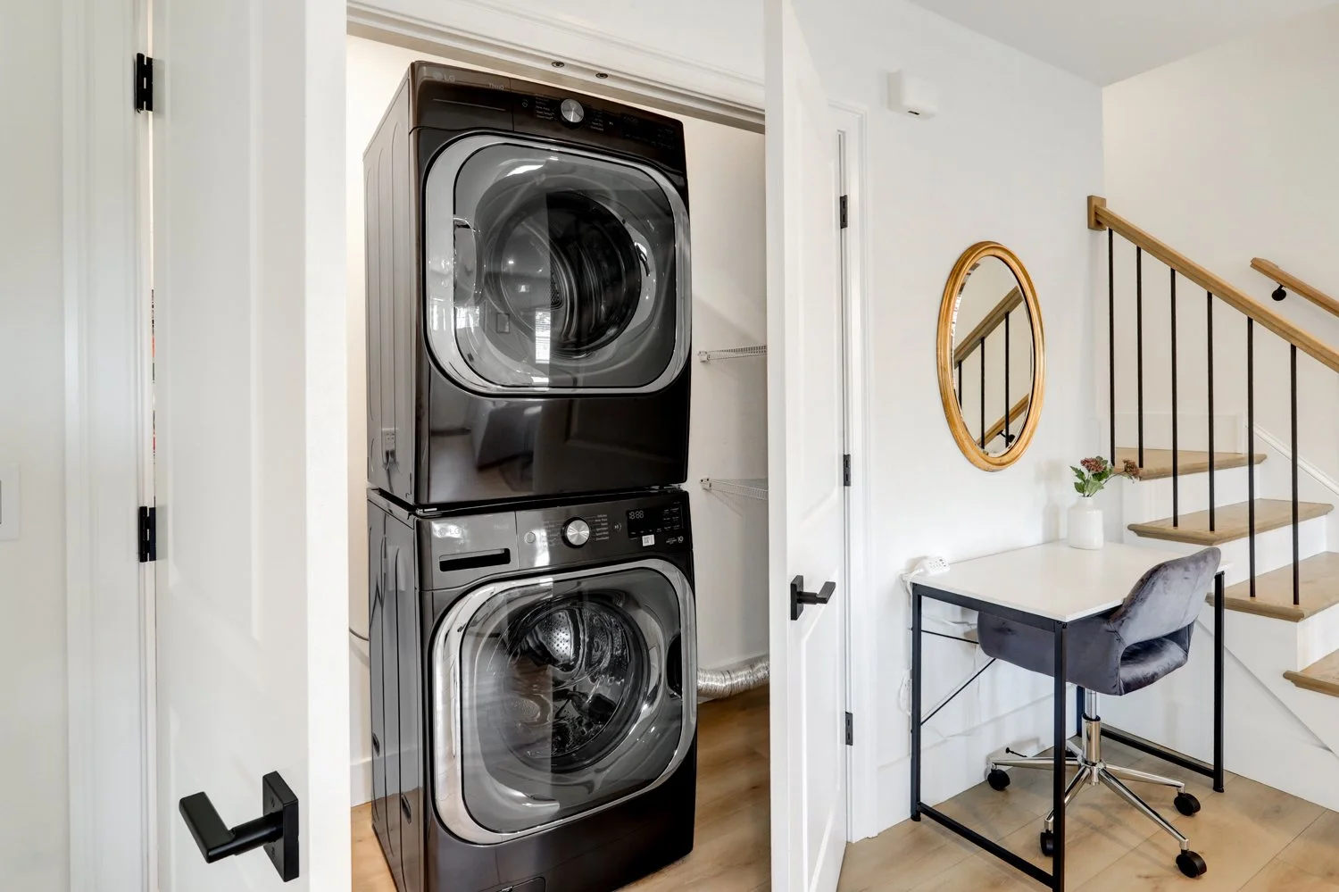 A laundry closet with stacked black washing machine and dryer, located next to a home office space with a desk, chair, and wall mirror, near stairs with wooden handrail.