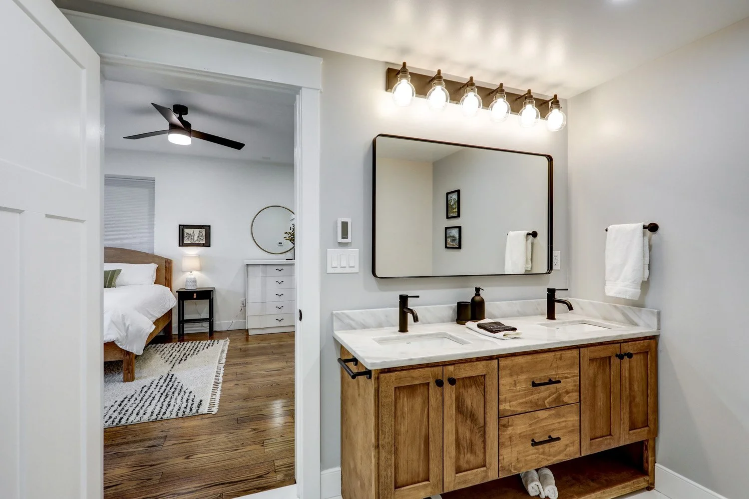 Bathroom with dual sinks, wood vanity, large mirror, and vanity lights. Bedroom with bed, nightstand, lamp, and decor visible through open door.