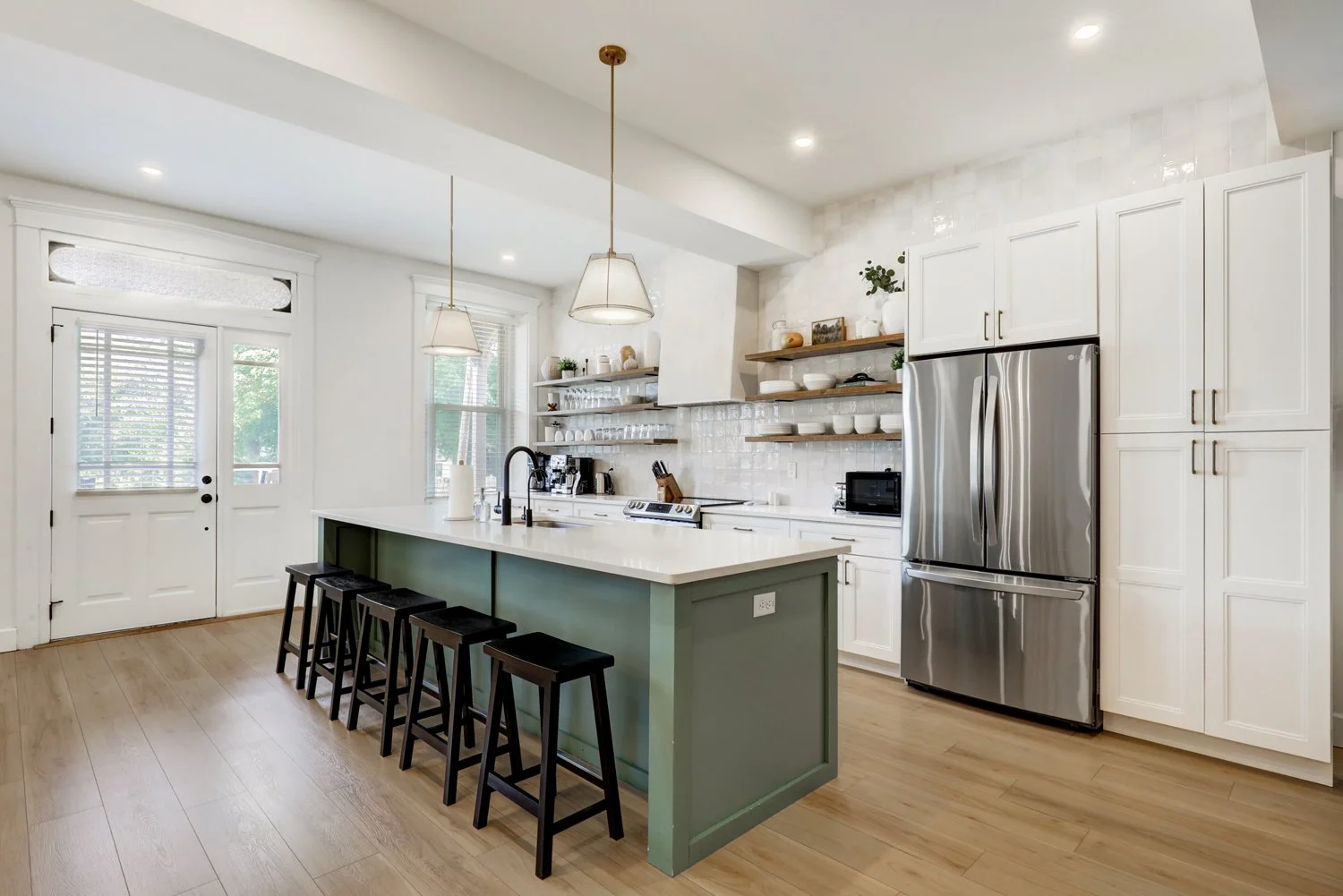 Bright kitchen with white cabinets, stainless steel refrigerator, open shelves with dishes, kitchen island with black stools, hardwood floors, and large windows.