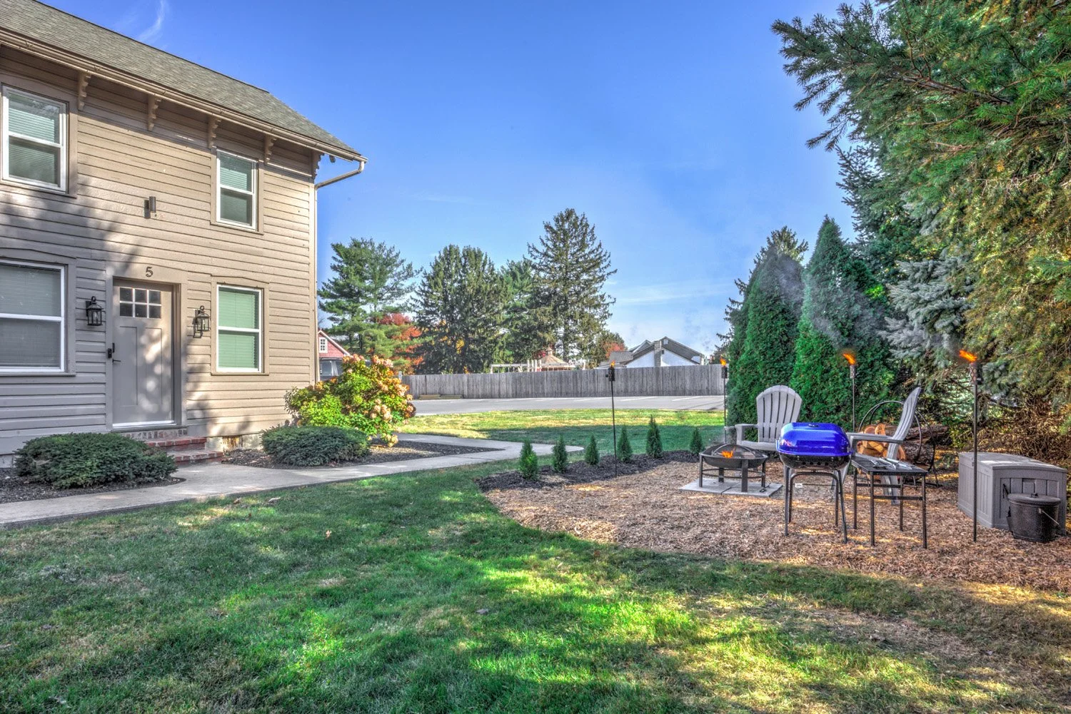 Backyard with patio area featuring chairs, a small table, a grill, and tiki torches, with a house on the left, a fence in the background, and trees surrounding the yard.