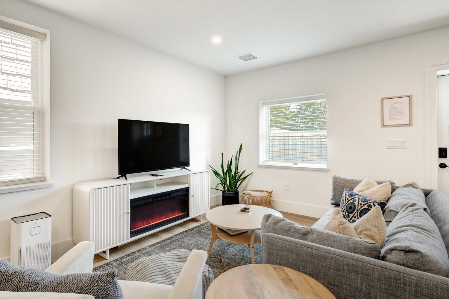 Living room with a gray sofa, a wooden coffee table, a black flat-screen TV on a white stand, a potted plant, and windows with blinds.