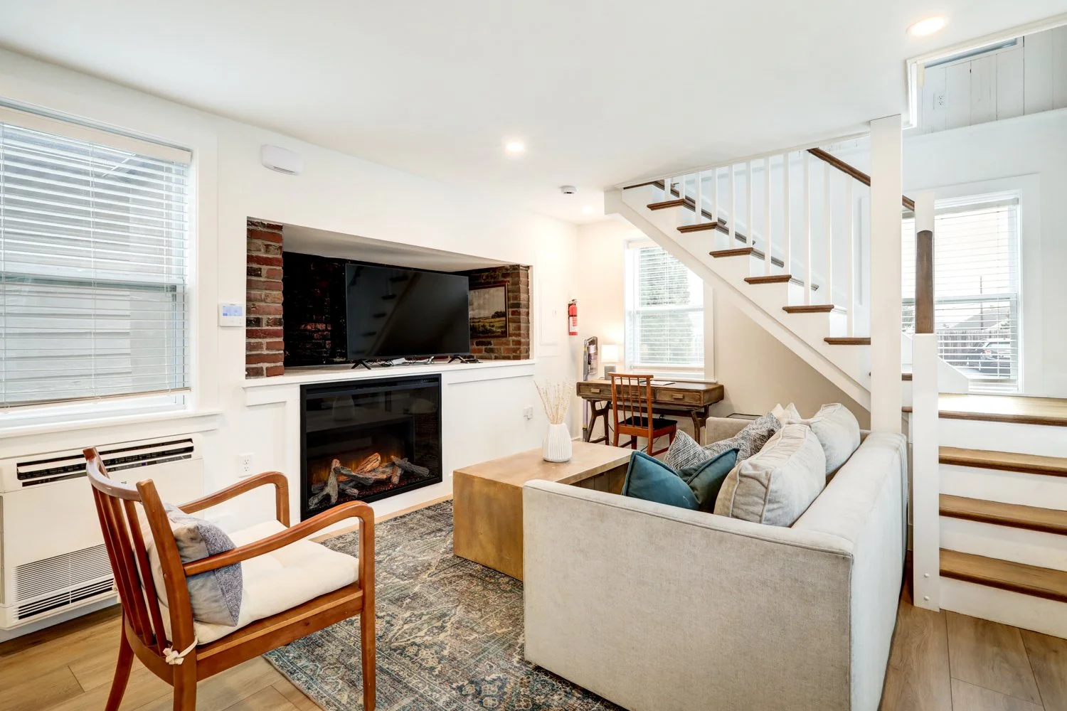 Bright living room with a beige sofa, wooden armchair, fireplace, flat-screen TV, staircase, and windows with blinds.