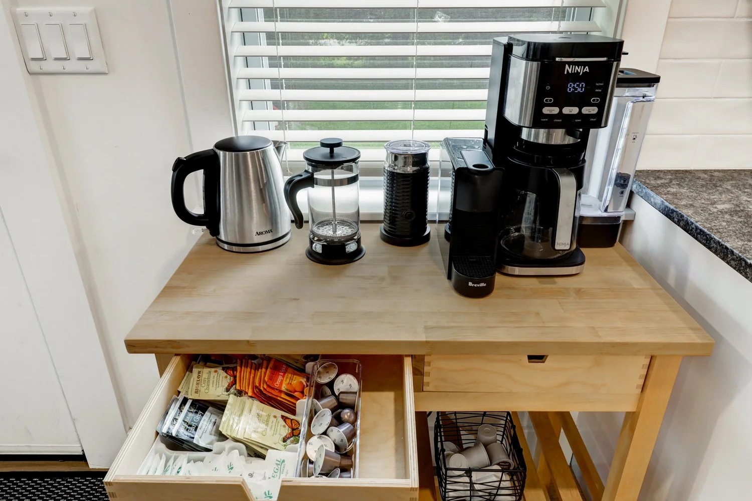 Coffee station on a wooden countertop with a kettle, French press, grinder, coffee maker, and water filter pitcher, next to a window with white blinds and a drawer containing tea, coffee, and tea bags, and a small basket with coffee pods.