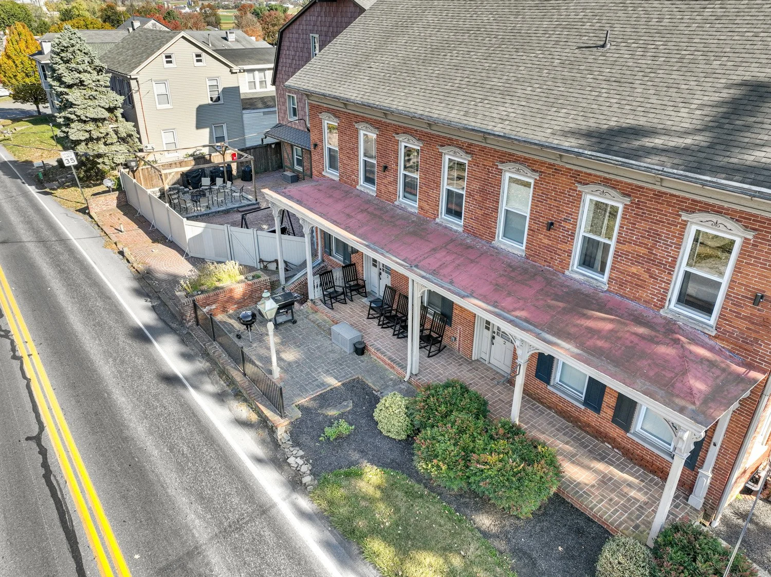 Aerial view of a two-story brick building with a front porch, outdoor seating area, and a backyard patio, situated along a street with yellow double lines and a sidewalk.