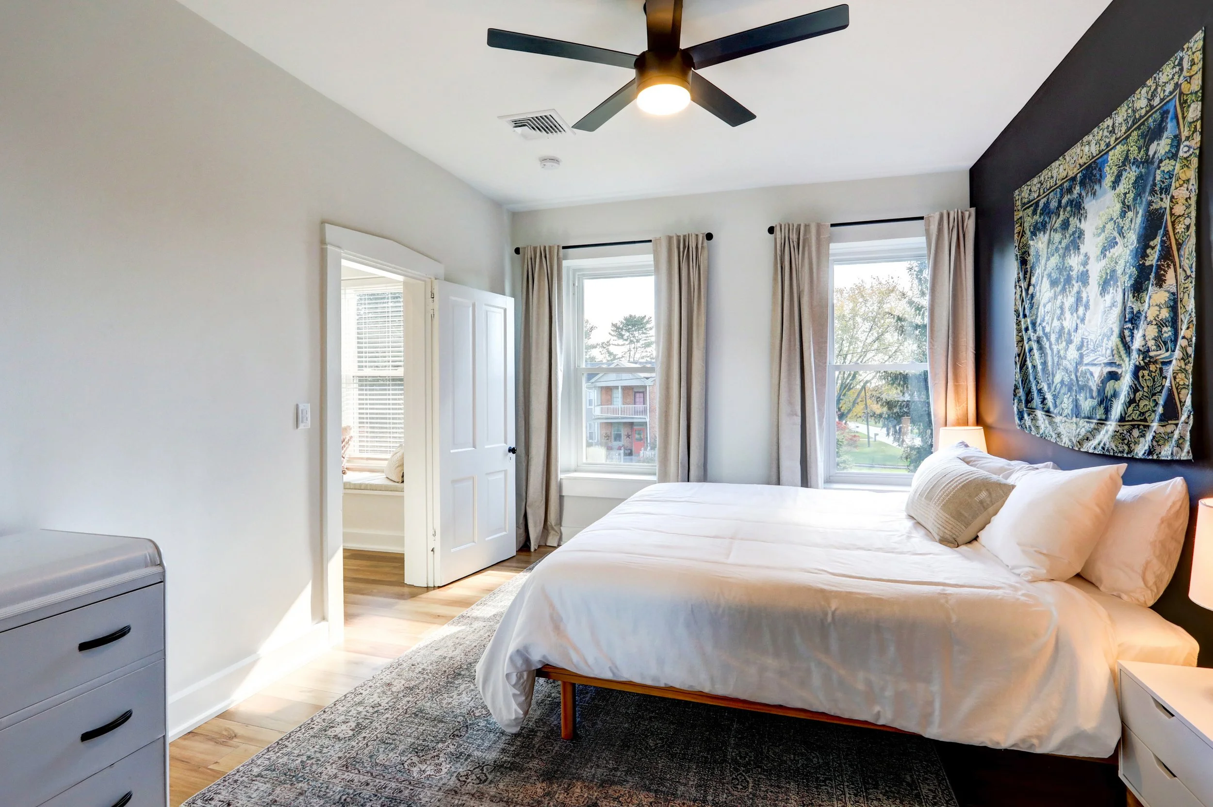A bedroom with white walls, three large windows with beige curtains, a bed with white bedding, a tapestry with a scenic landscape hanging on a dark wall, a nightstand with a lamp, a ceiling fan with lights, and a dresser with black handles.