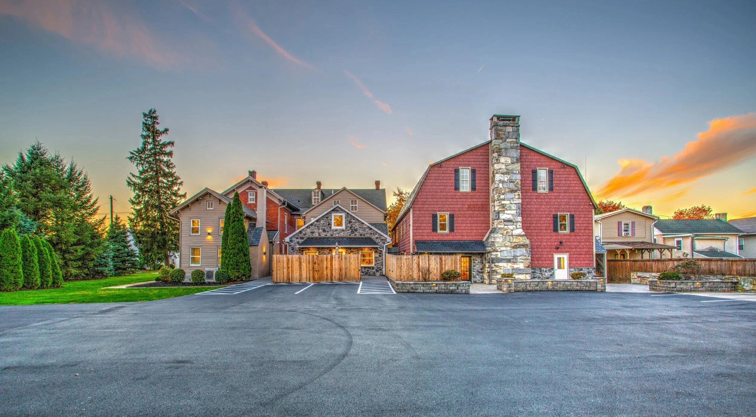 Photo of multiple houses in a neighborhood at sunset, with trees, a parking lot, and a colorful sky.