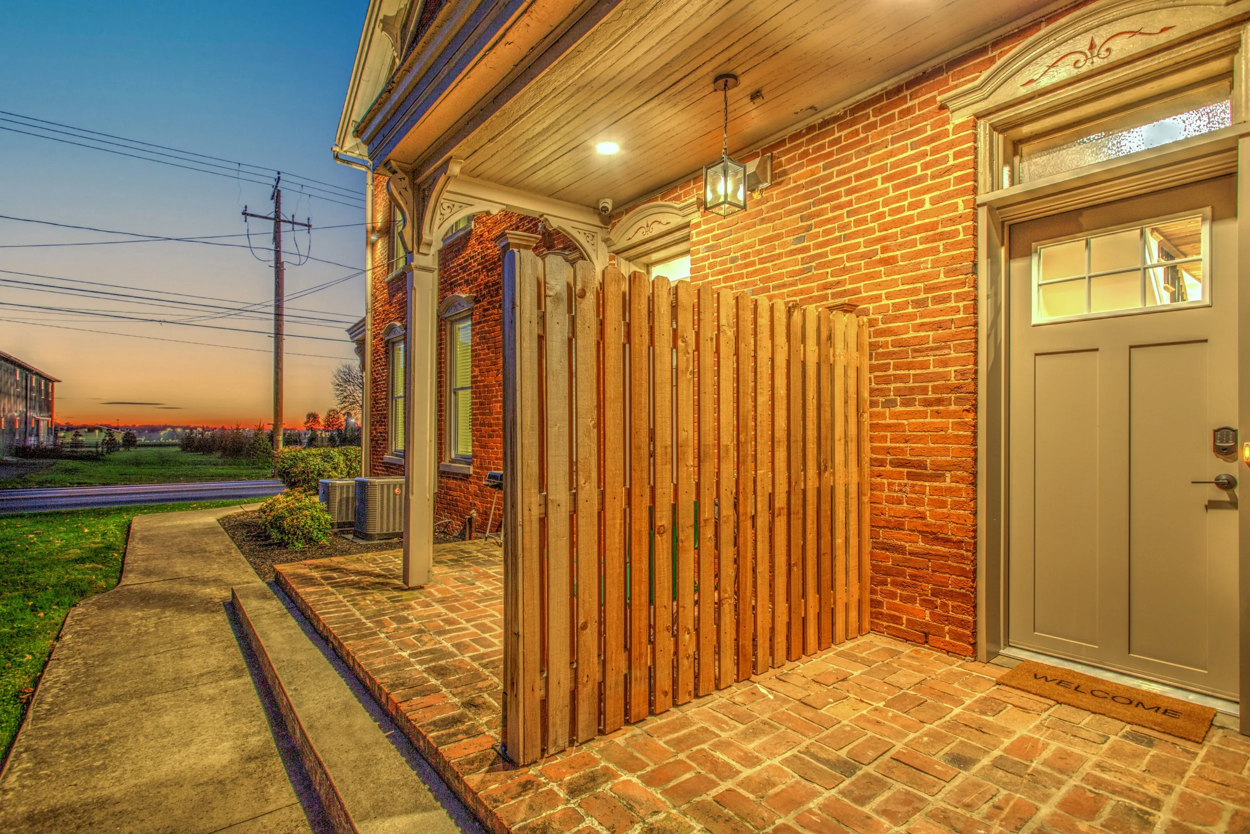 The image shows the exterior of a brick house during sunset with a porch area. The porch has a wooden privacy fence, a hanging lantern light fixture, and a white door with a small window. There is a welcome mat in front of the door. The background fe