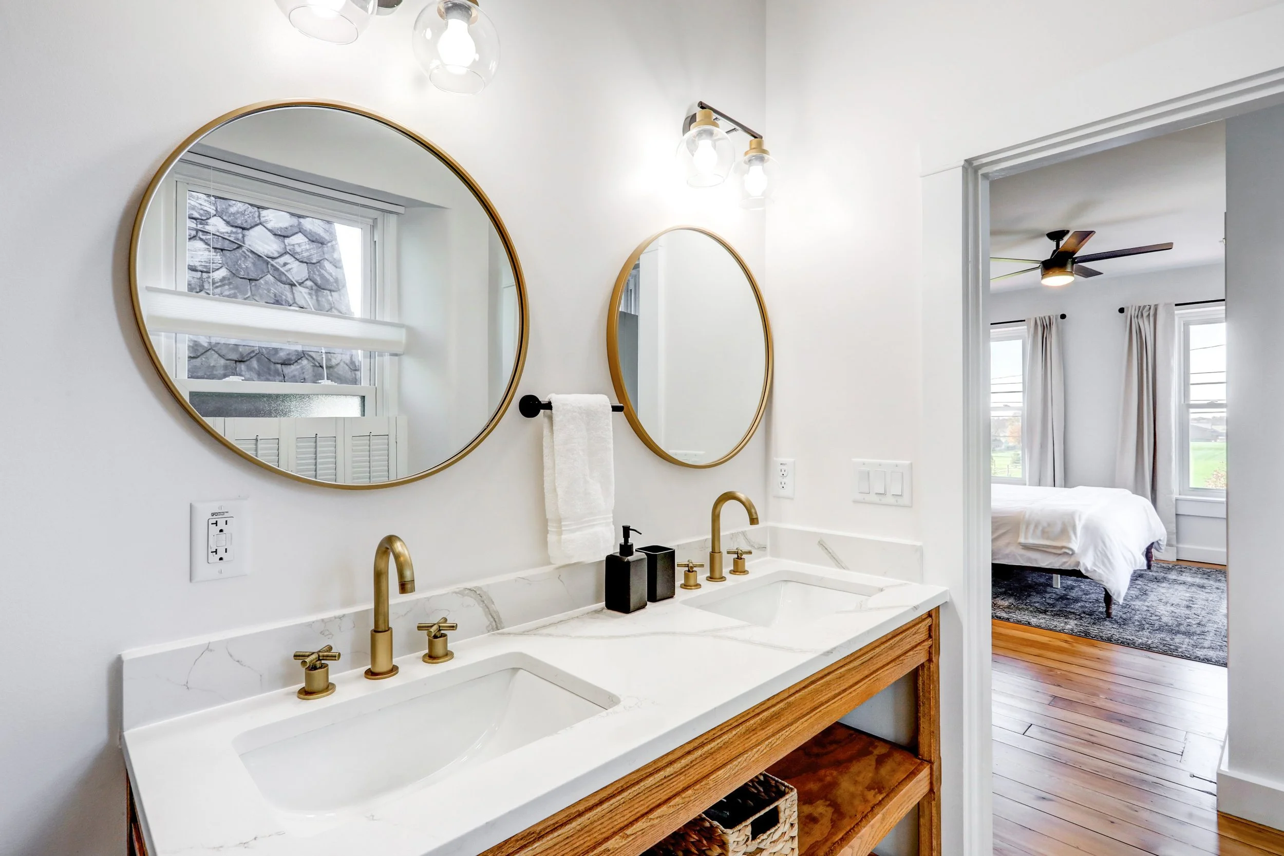 Bathroom vanity with double oval mirrors, brass faucets, and wooden cabinet below a marble countertop. Visible part of a bedroom with a bed and ceiling fan seen through the doorway.
