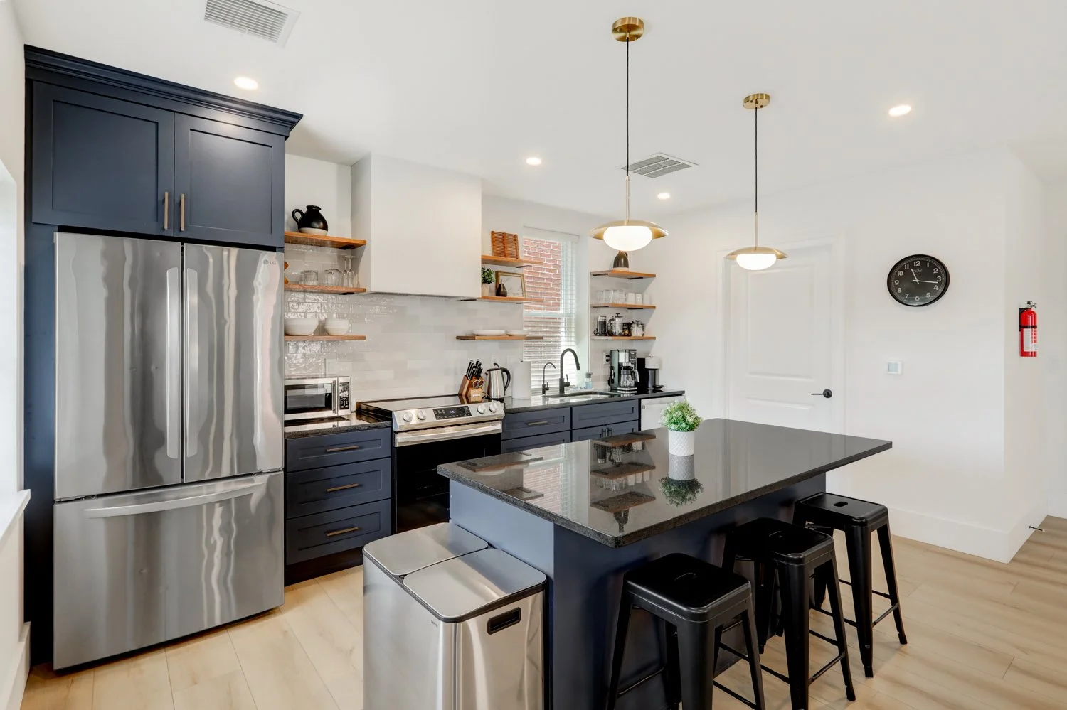 Modern kitchen with black cabinetry, stainless steel appliances, open wooden shelves, white backsplash, black countertop island with barstools, white walls, natural light from window, pendant lights, wall clock, fire extinguisher.