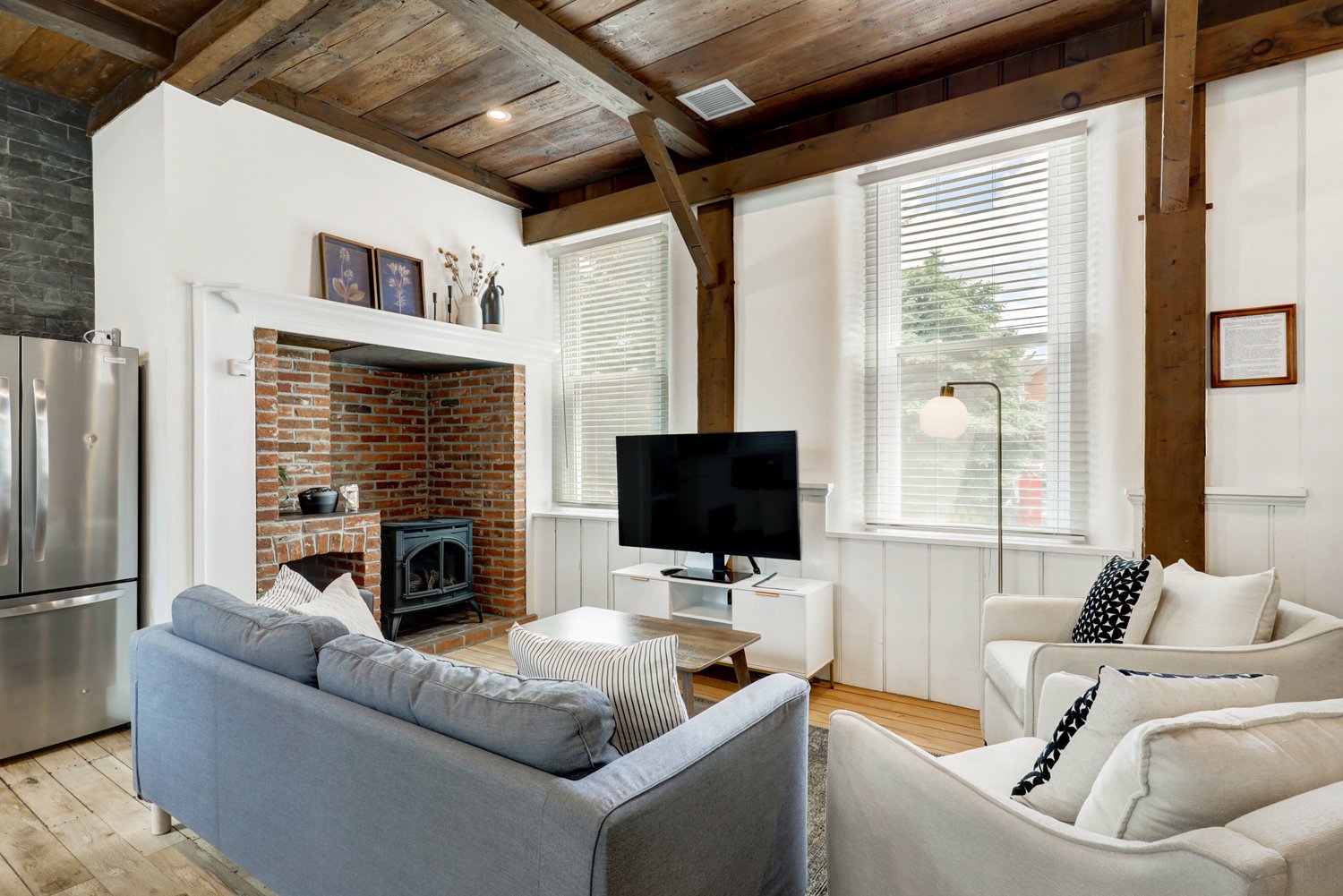 Living room with white walls, large windows with blinds, a brick fireplace, and wooden ceiling beams. Contains a gray sofa, white armchairs, a television, and a small coffee table.