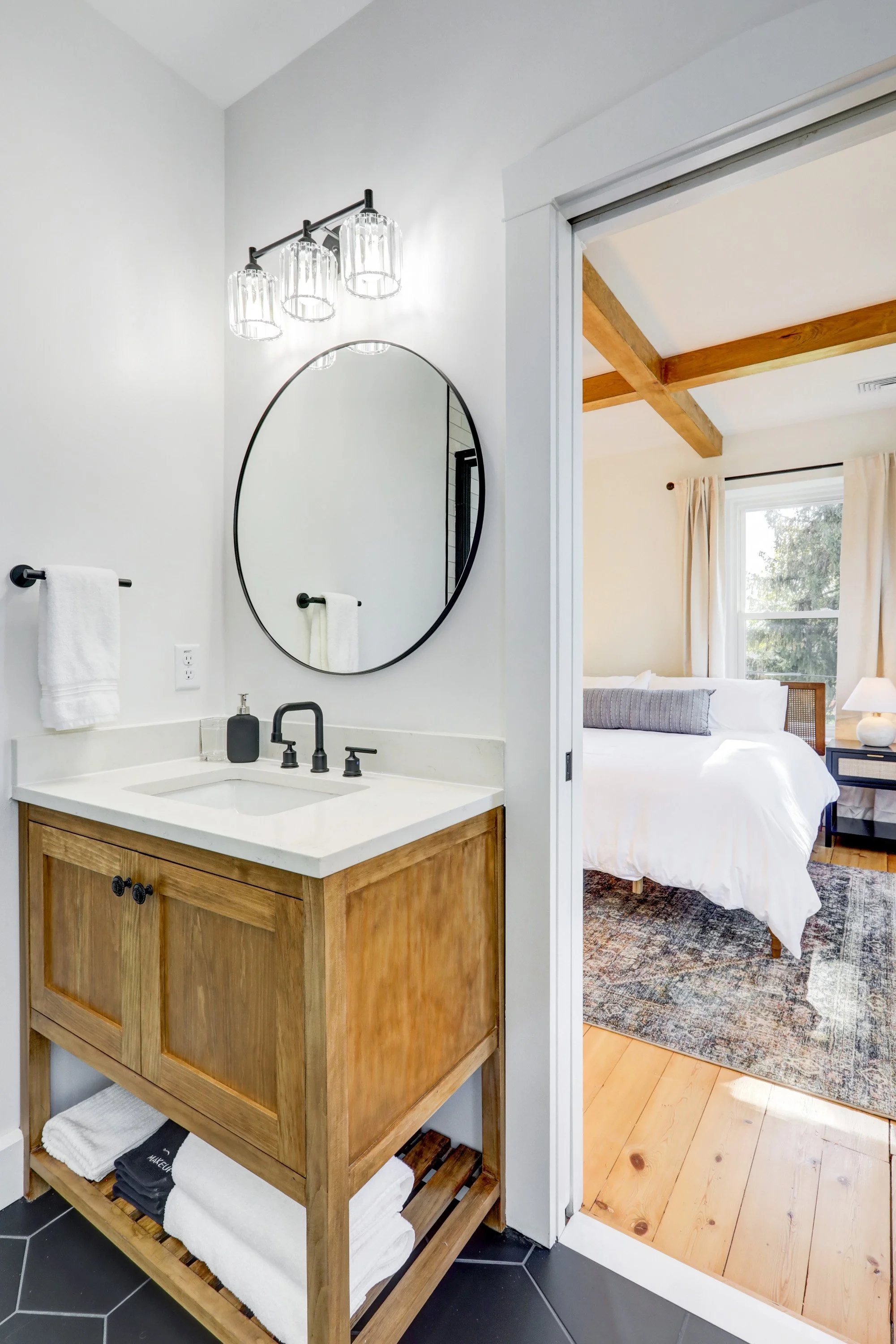 Bathroom vanity with a wooden cabinet, black faucet, and round mirror. Visible towels and soap dispenser. Adjacent bedroom with a bed, window, and exposed wooden beams.