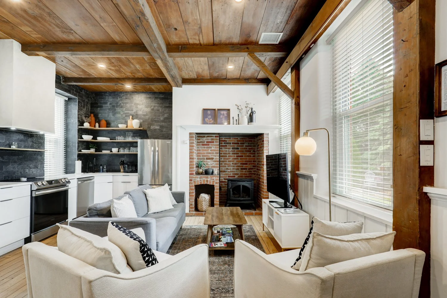 Living room with a wood-beamed ceiling, brick fireplace, white cabinetry, black brick wall in the kitchen area, gray sofa, white armchair, coffee table with magazines, TV on a white stand, large windows with blinds, and decorative items on shelves.