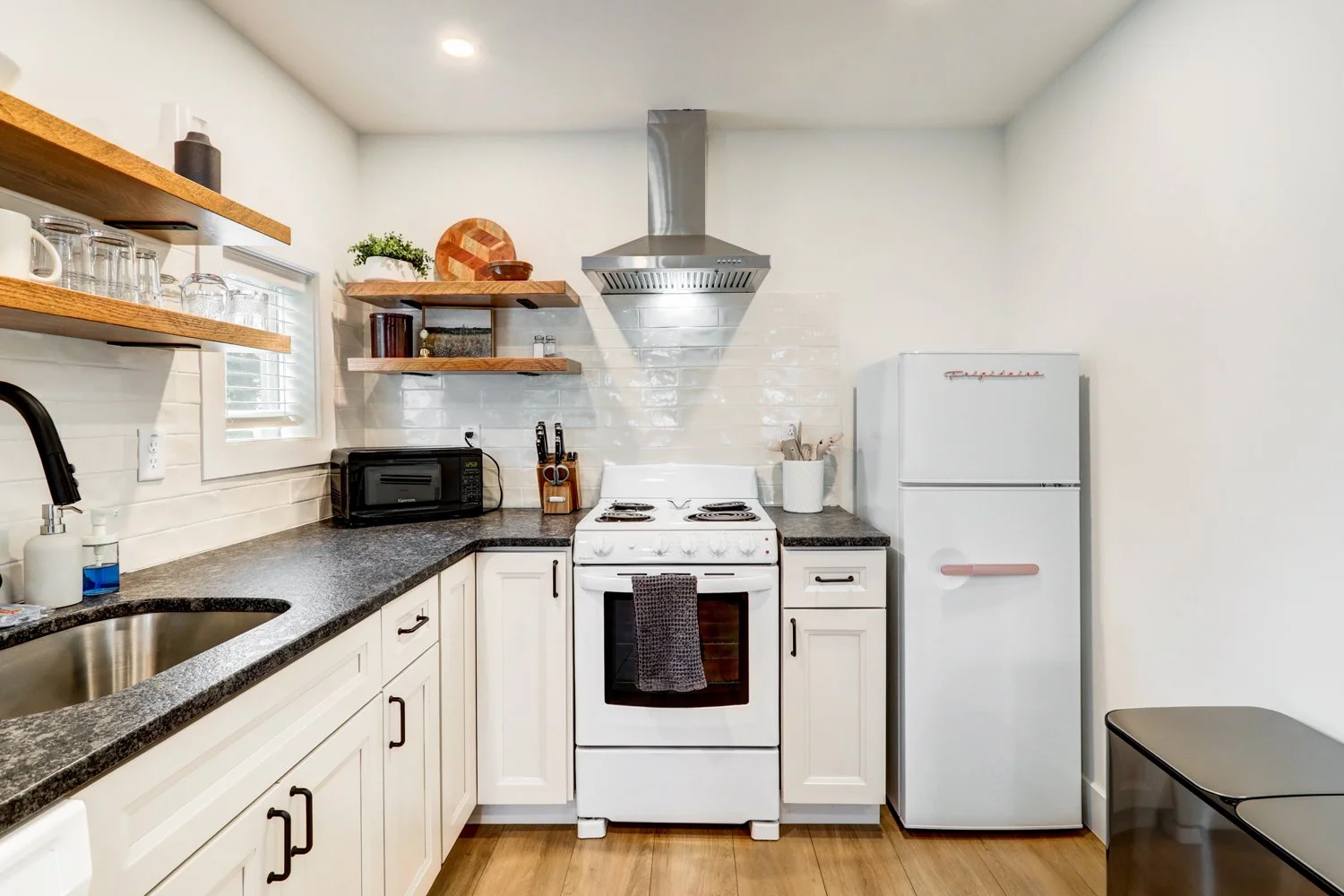 Modern kitchen with white cabinets, black countertop, stainless steel sink, open wooden shelves with glassware, black microwave, white stove with a towel hanging, white refrigerator with pink handle, wooden flooring, small window with blinds.