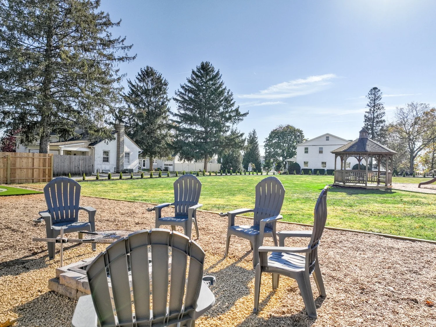 An outdoor scene with six gray Adirondack chairs arranged around a fire pit on a gravel surface. In the background, there is a well-maintained lawn, a gazebo, and several houses and tall trees under a clear blue sky.