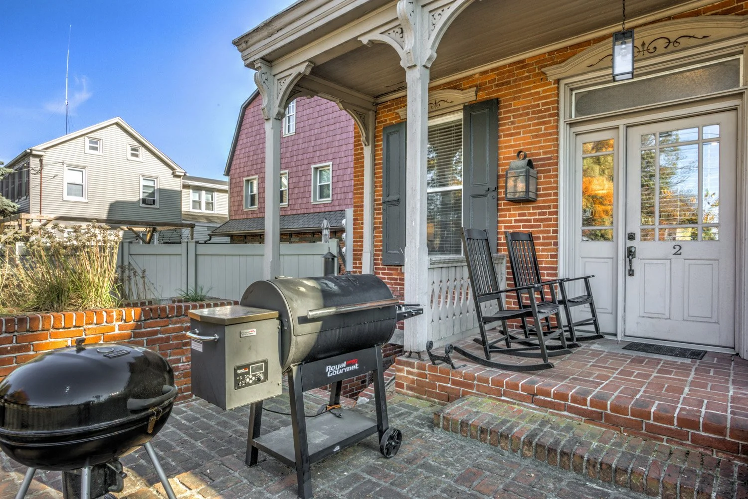 Front porch with two black rocking chairs, a grill, and a small black round smoker, brick flooring, brick house with white door labeled '2', black window shutters, and a lantern-style light fixture.