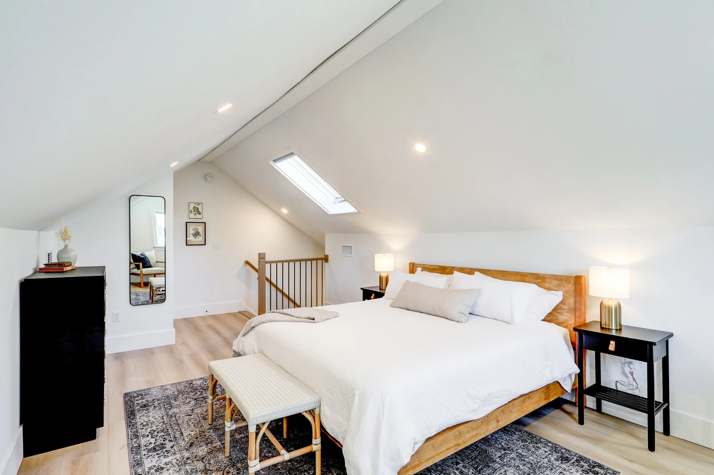 A spacious bedroom with two beds, a wooden headboard, nightstands with lamps, a bench at the foot of the bed, a mirror, and wall art, with a skylight and sloped ceiling.