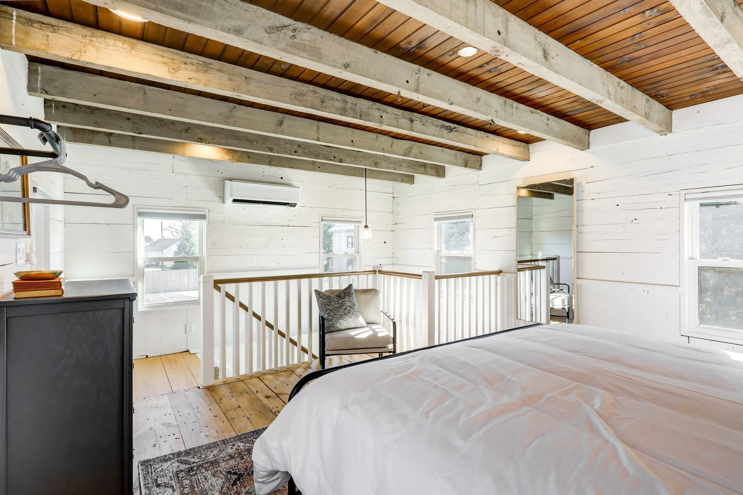 A bright bedroom with white wooden walls, a wooden ceiling with exposed beams, multiple windows, and a small seating area with a cushioned chair and a mirror. There is an air conditioning unit on the wall.
