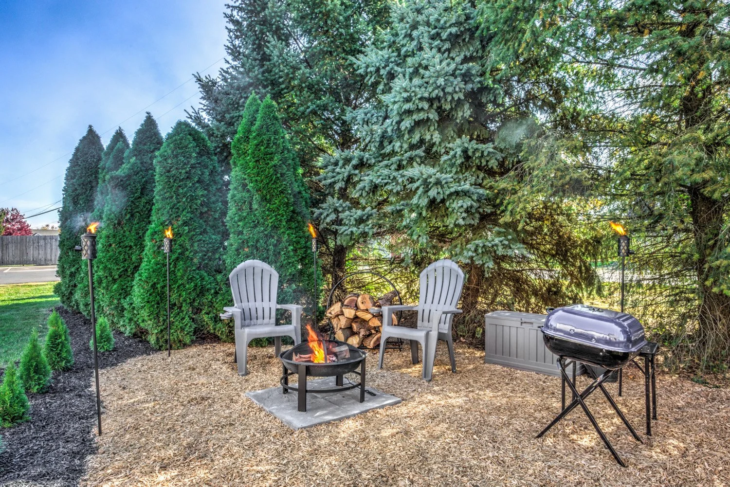 Outdoor backyard patio with a fire pit surrounded by two Adirondack chairs, a stack of firewood, a barbecue grill, and tiki torches, with tall evergreen trees and a clear sky in the background.