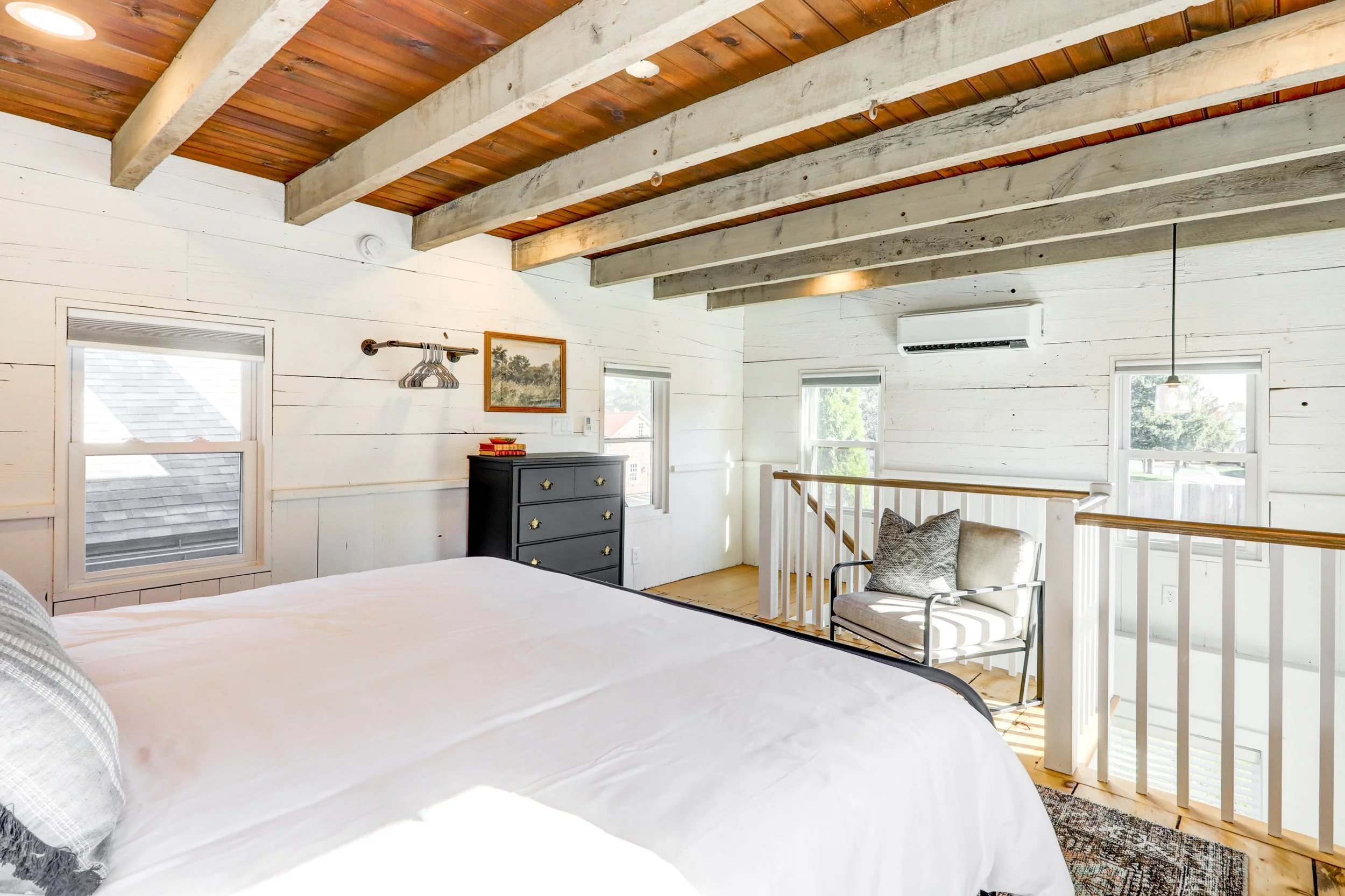 Bright top-floor bedroom with white wooden walls and ceiling beams, a window, a black dresser, a cozy sitting nook with a chair, and wood flooring.
