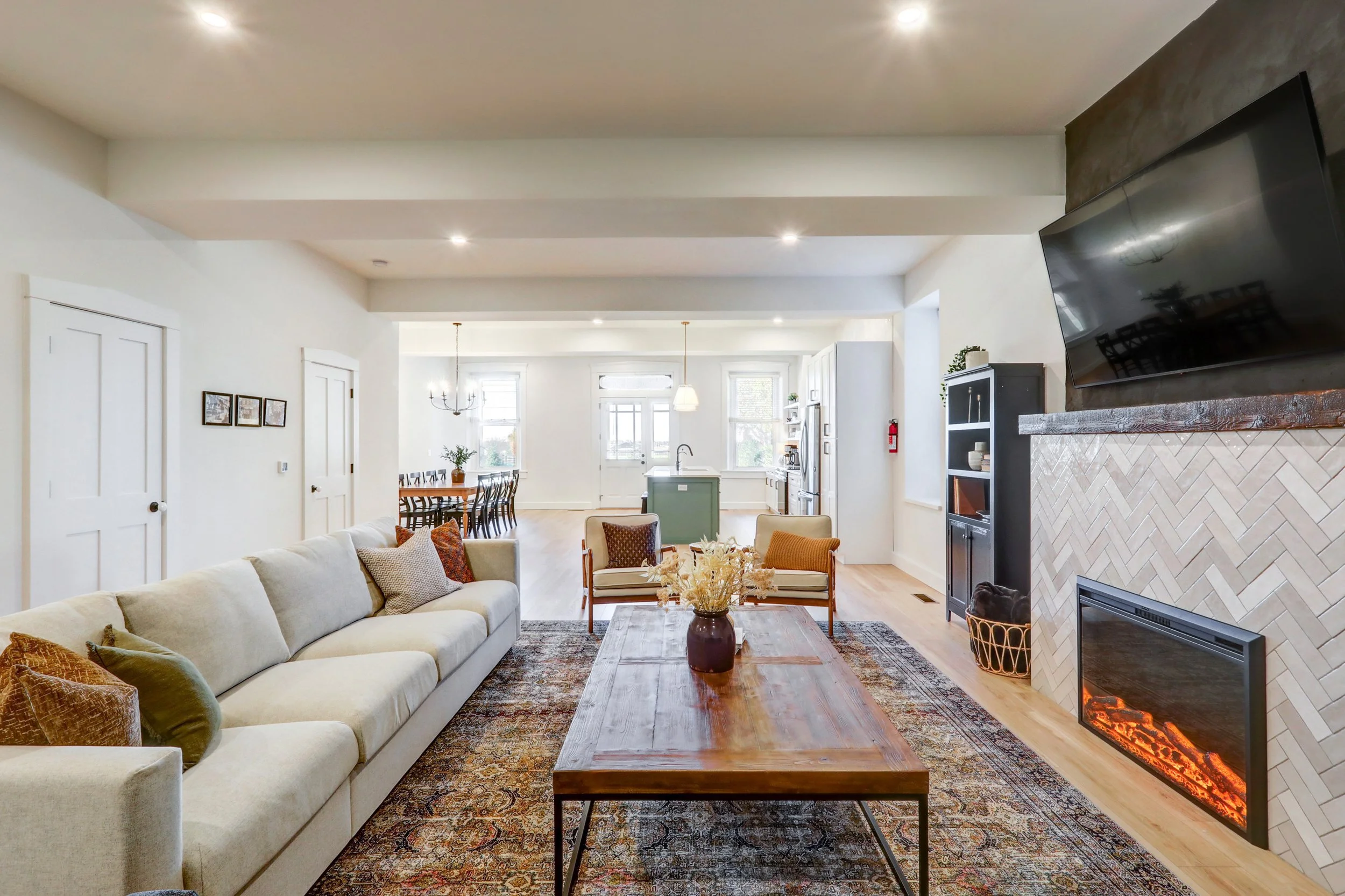 Living room with a cream-colored sofa, wooden coffee table, patterned area rug, and a fireplace with a herringbone tile design. Background includes a dining area, kitchen with island, and large windows bringing in natural light.