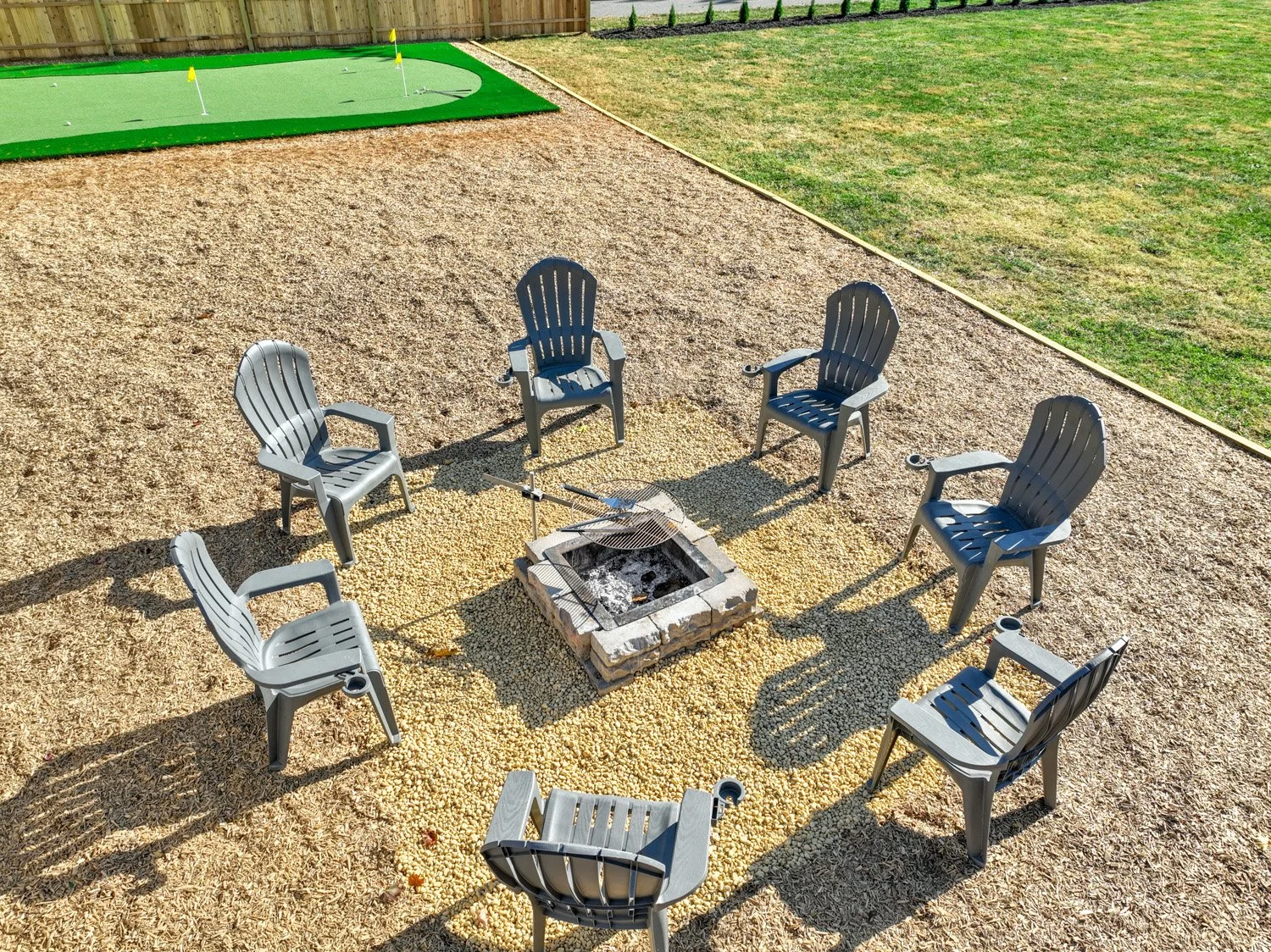 Six outdoor gray Adirondack-style chairs arranged around a fire pit in a backyard with a gravel ground, green lawn, and a small golf putting green in the background.