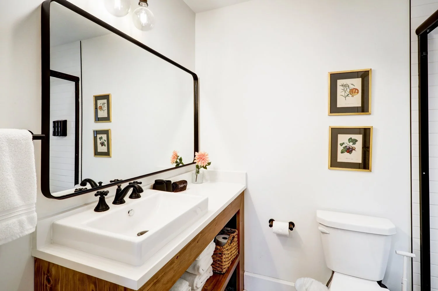 A bathroom with a large mirror, a white sink with black fixtures, a wooden vanity with towels underneath, a small basket, pink flowers in a glass vase, framed botanical prints on the white wall, and a toilet.