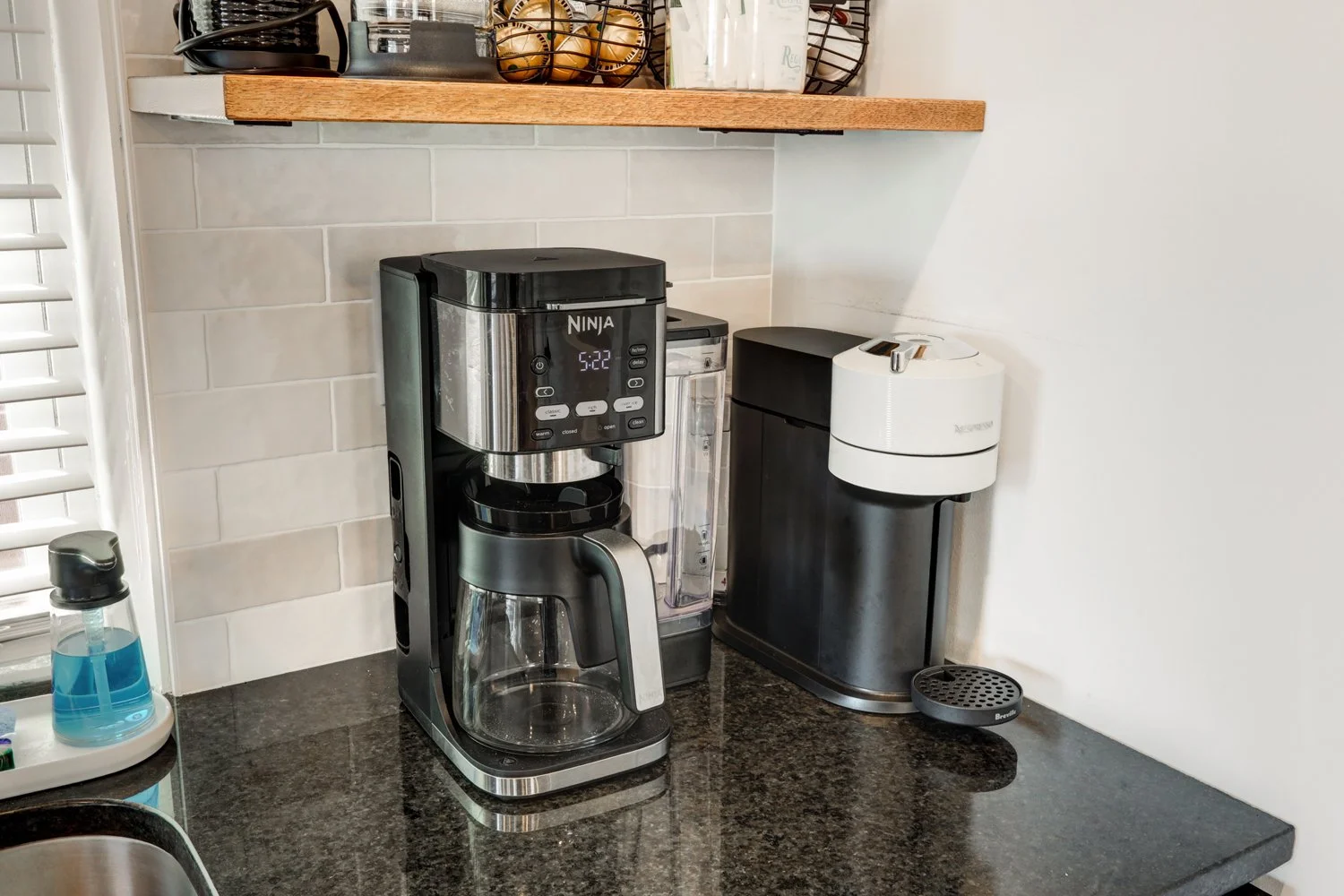Coffee maker, water filter, and bread maker on a black granite kitchen countertop with a window and white backsplash tiles.