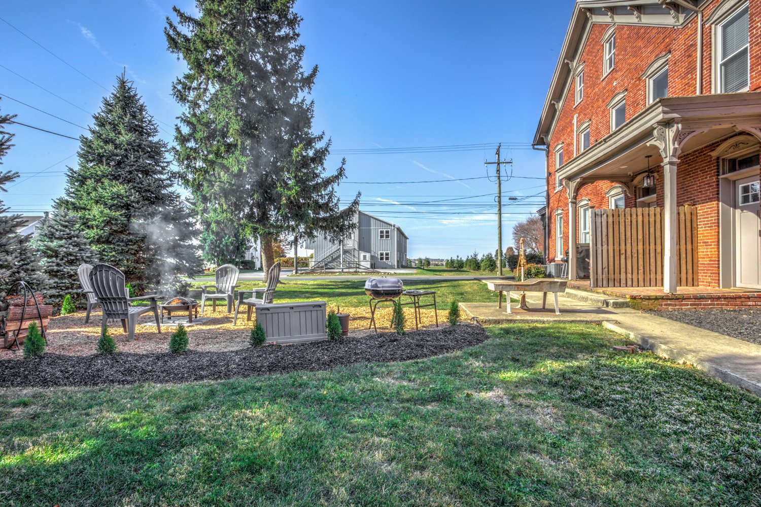 Backyard with Adirondack chairs around a fire pit, presence of a grill, a fenced area, mature trees, well-maintained lawn, and a brick house on the right side, under a clear blue sky.