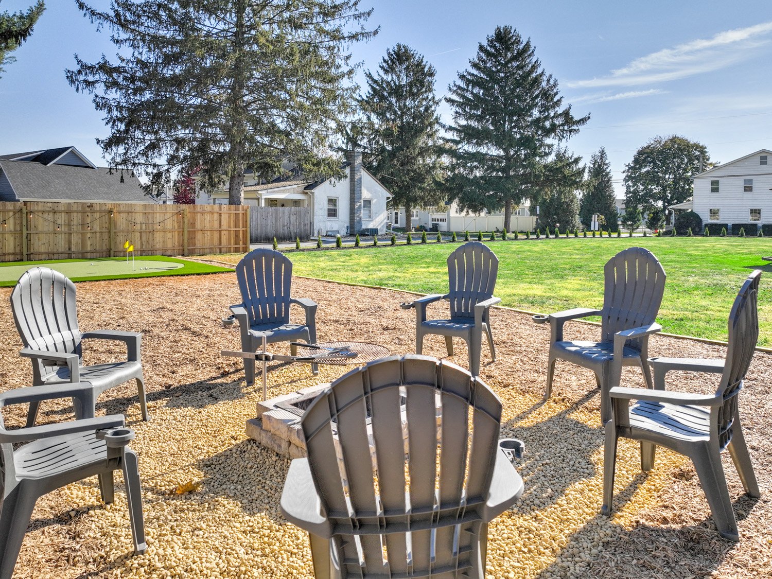 Backyard with outdoor chairs arranged around a fire pit on gravel, green lawn, tall trees, and houses in the background on a sunny day.