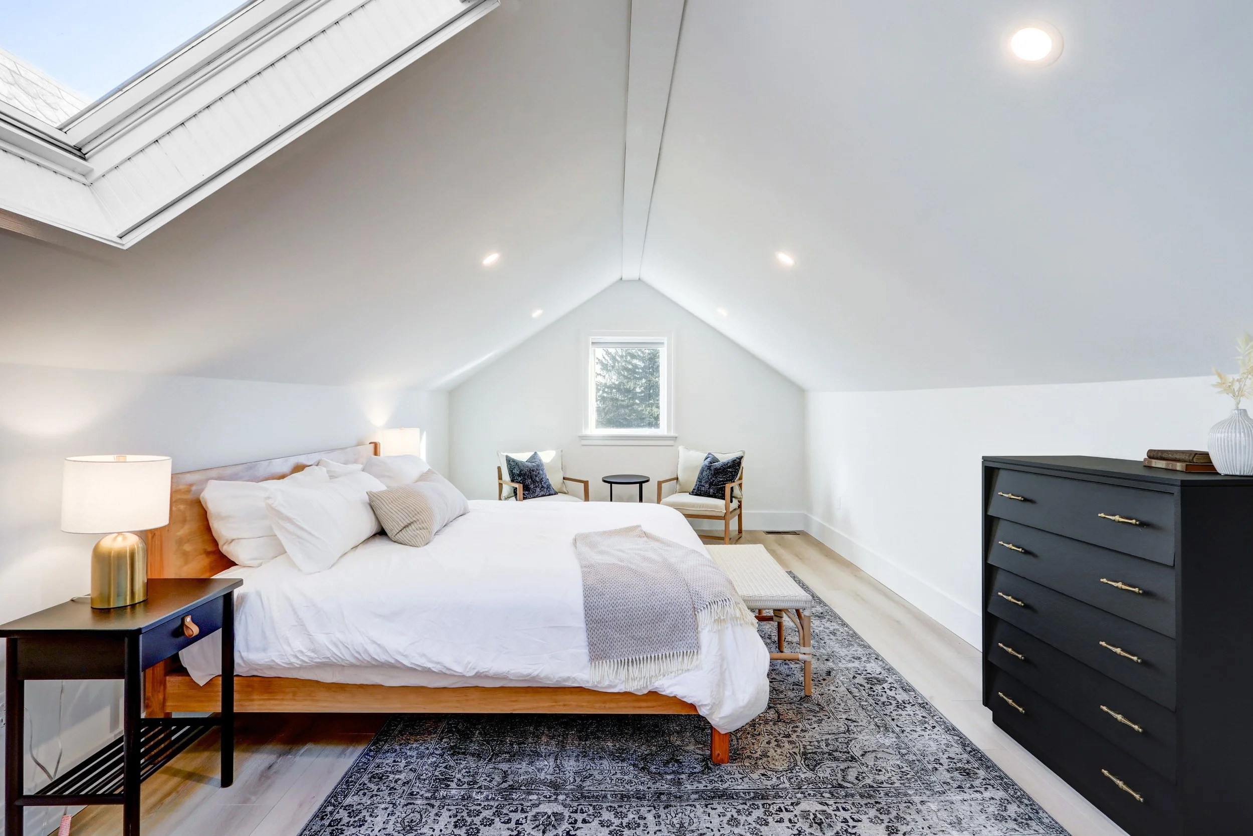 A bedroom with a sloped ceiling, a bed with white bedding and pillows, a bedside table with a gold lamp, a black dresser, a window, and two chairs with pillows.