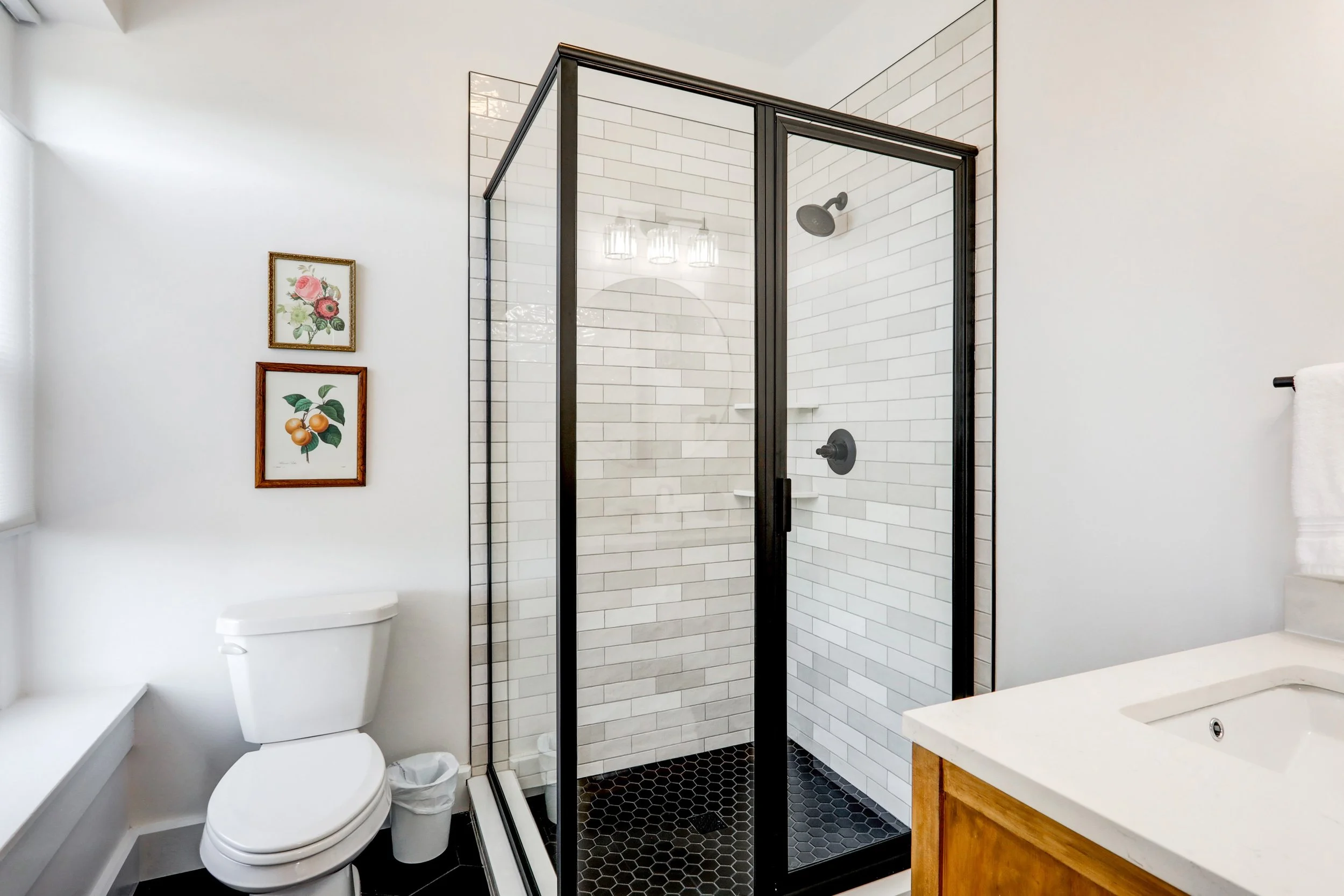 Modern bathroom with glass-enclosed shower with white subway tiles, black fixtures, a white toilet, and a wooden vanity with a white countertop. There are two framed floral art pieces on the white wall.