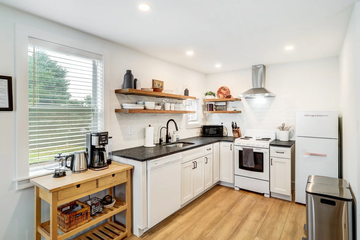 Kitchen with white cabinets, black countertops, wooden floating shelves, a window with blinds, and various appliances including a coffee maker, toaster, microwave, oven, and refrigerator.