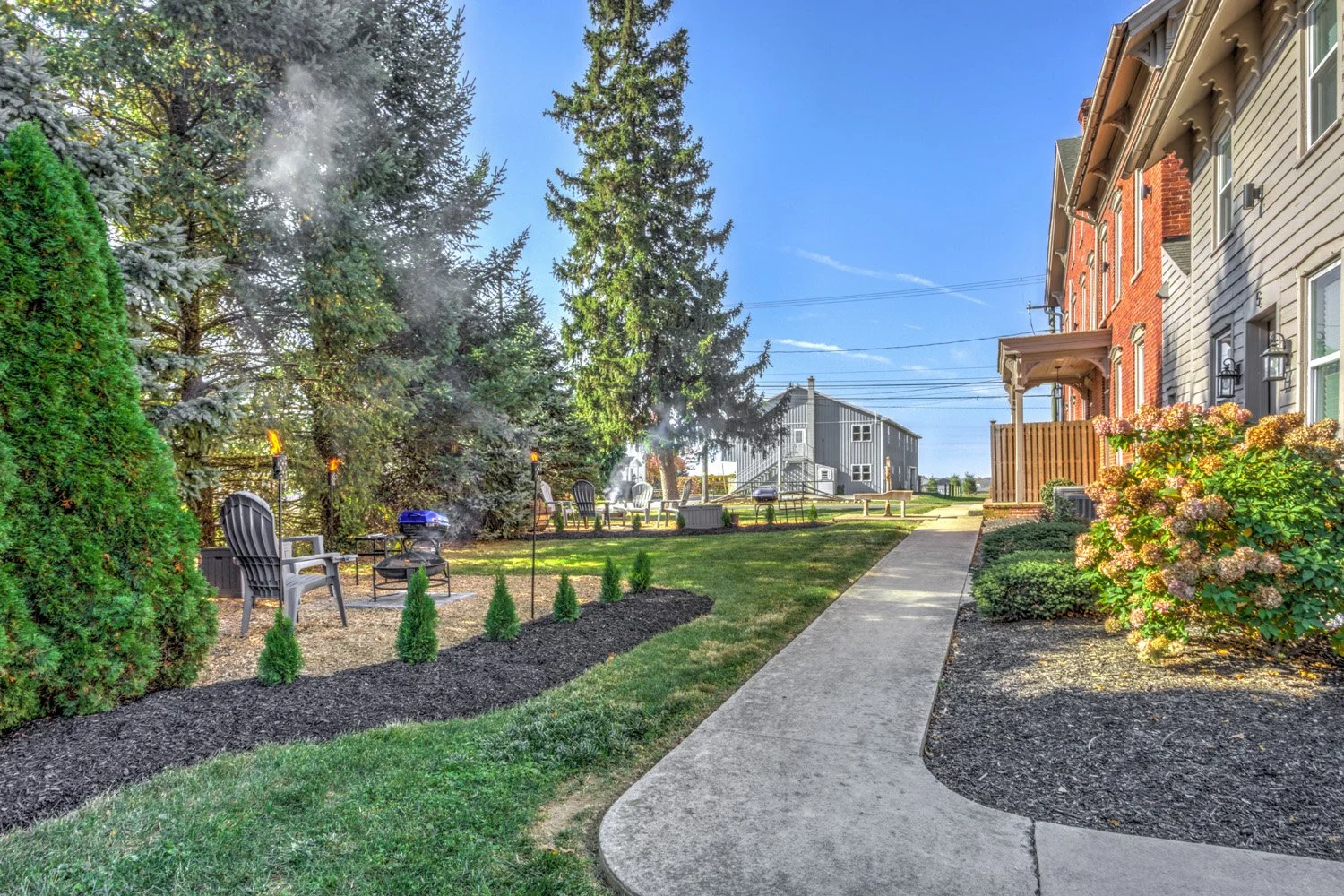 Backyard with a concrete path leading to a lawn with outdoor seating, surrounded by trees, shrubs, and a fire pit, adjacent to a multi-story house with a small porch and flowering bushes.