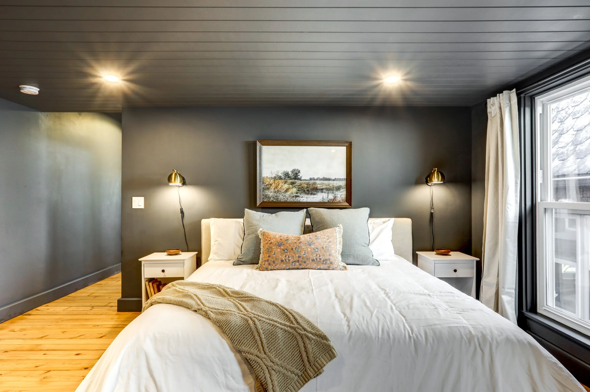 A cozy bedroom with a large bed, gray walls, two white nightstands, and a landscape painting above the headboard. The room features recessed ceiling lights and a large window with white curtains, showing part of a sloped roof outside.