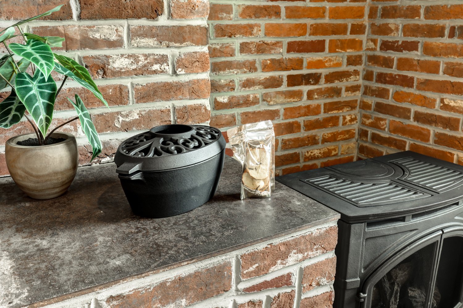 A house interior corner with a brick wall, a plant in a beige pot, a black decorative diffuser, a small bag of burnt wood or essential oils, and a wood stove with a metal grate on top.