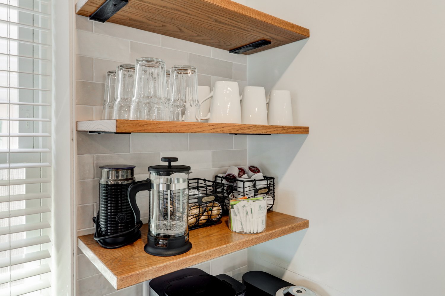 Kitchen shelves with glasses, cups, coffee maker, French press, coffee capsules, and sugar packets.