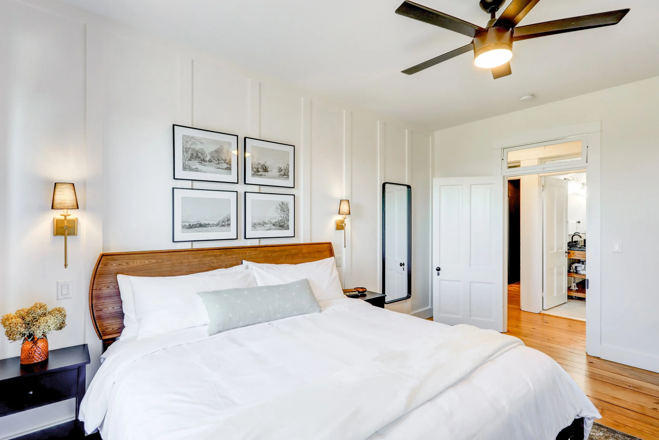 A bedroom with a bed, nightstands, lamps, and overhead fan, featuring white walls, framed artwork, and a door leading to another room.